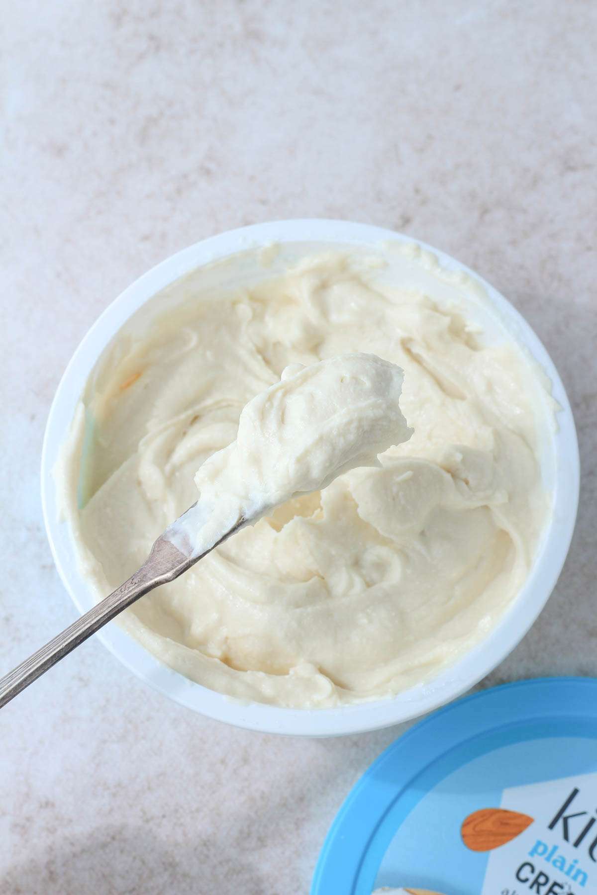 An open tub of dairy-free cream cheese with a spreading knife on a tan counter.
