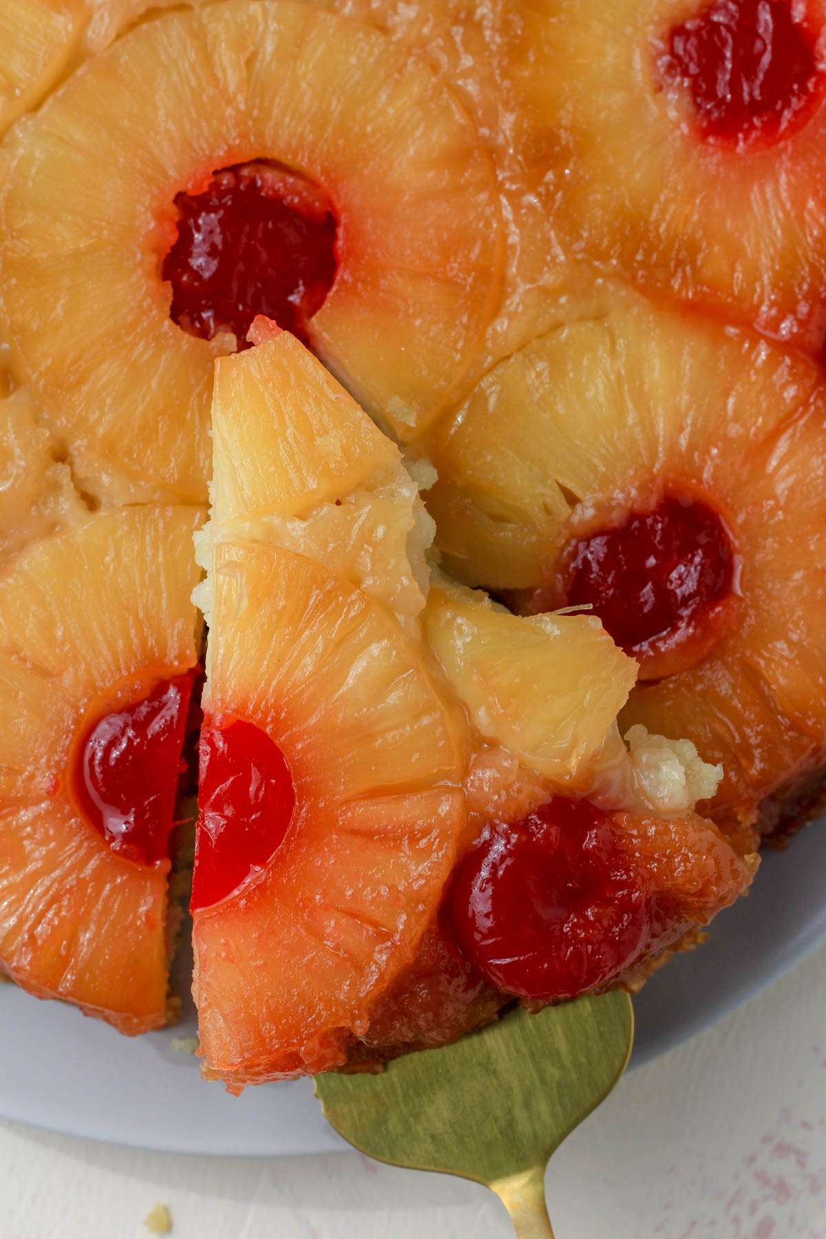 A close up wedge of vegan pineapple upside down cake on a cake plate with a gold server pulling a slice out.