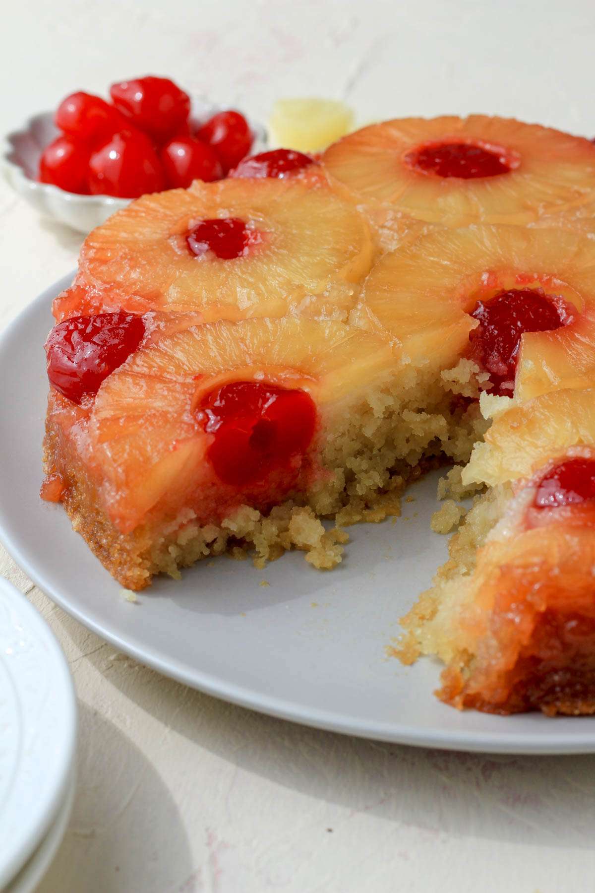 From the side, the left half of a pineapple upside down cake with a wedge missing from the cake plate and a small dish of cherries in the back left.