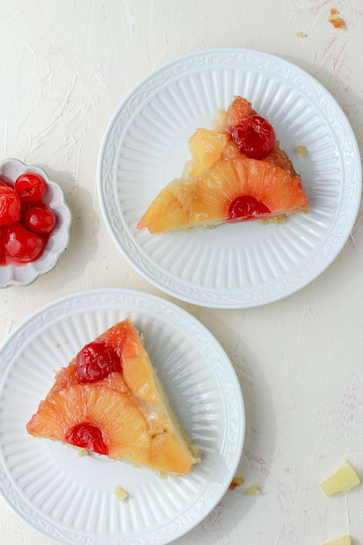Two small white plates with wedges of vegan pineapple upside down cake with a small dish of cherries in the middle left.