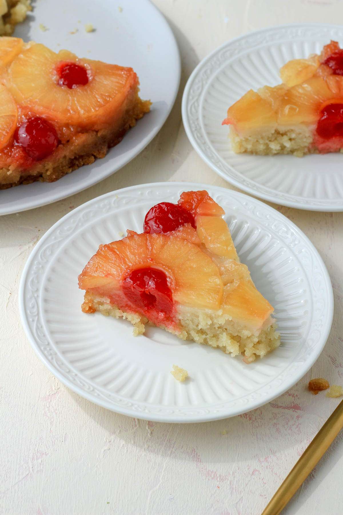 From the side, a small white plate with a slice of pineapple upside down cake with a white plate in the back right with another slice of cake and a cake plate in the back left.