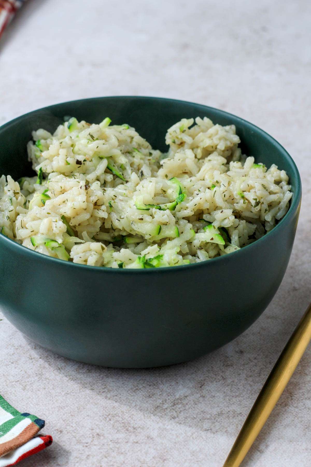 A green bowl on a cream counter filled with seasoned rice and zucchini with a gold fork to the right.