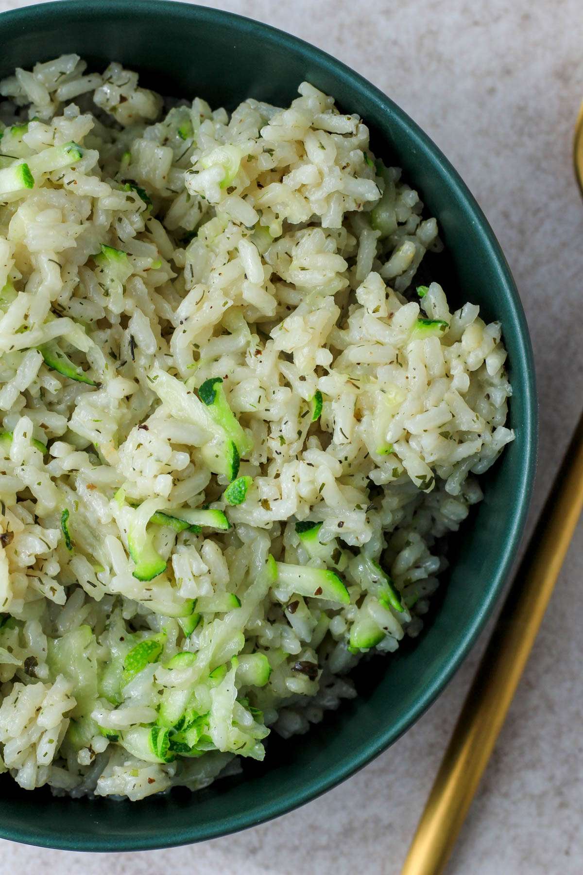 A top down image of a green bowl filled with vegan seasoned zucchini rice with a gold fork to the right.