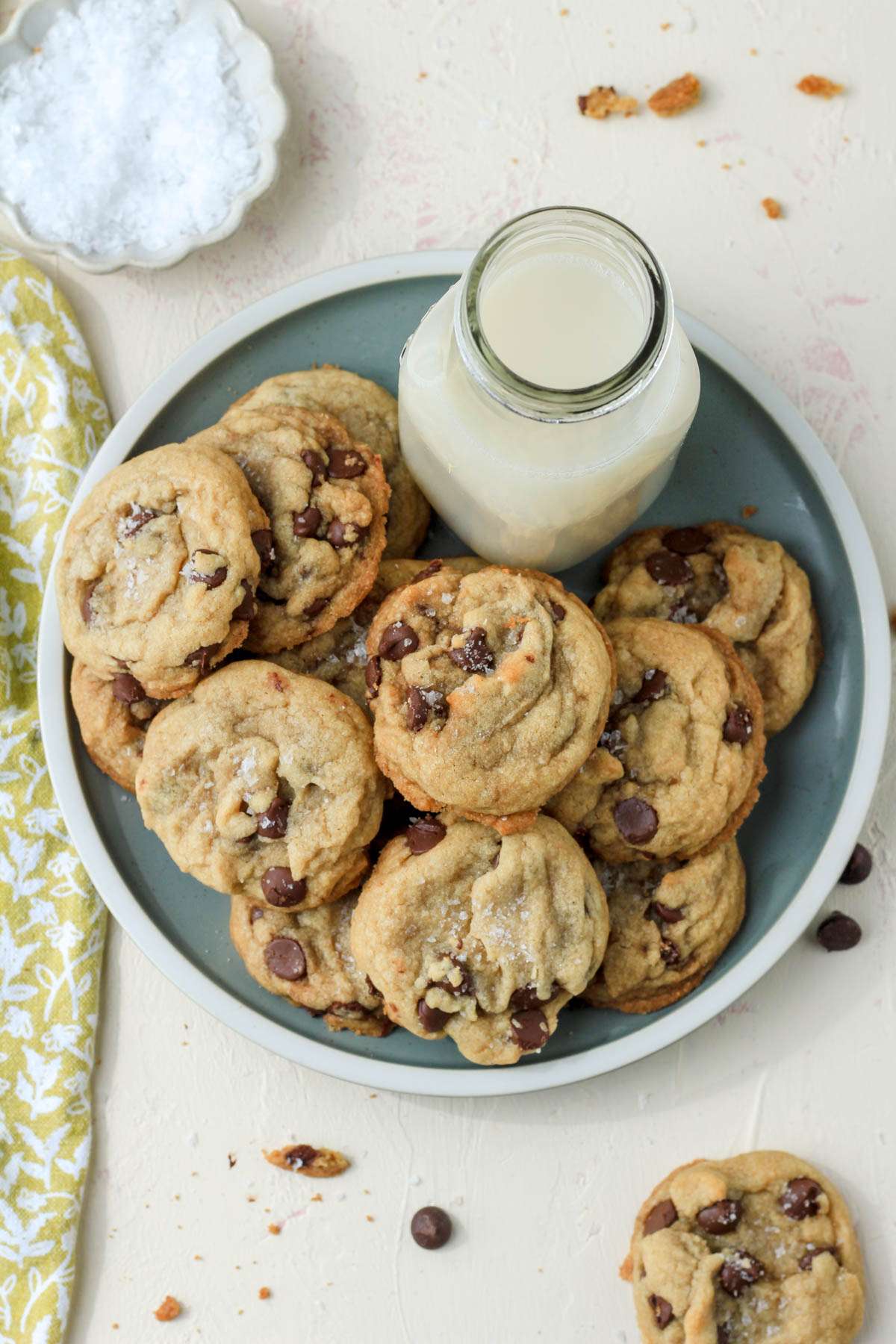 A top down image of a green plate with a cream rim topped with dairy-free chocolate chip coconut oil cookies with a non-dairy jug of milk on the plate and a pinch bowl of flaky sea salt in the top left.