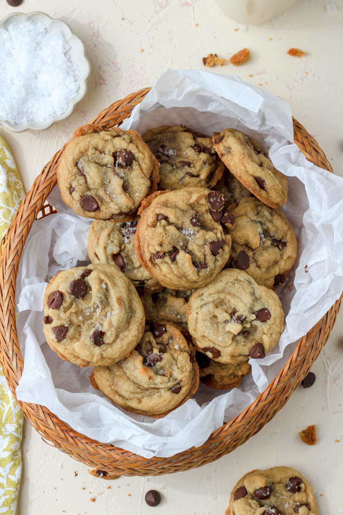 A top down image of a brown basket lined with parchment paper and filled with coconut oil chocolate chip cookies with a small pinch bowl of flaky sea salt in the top left.