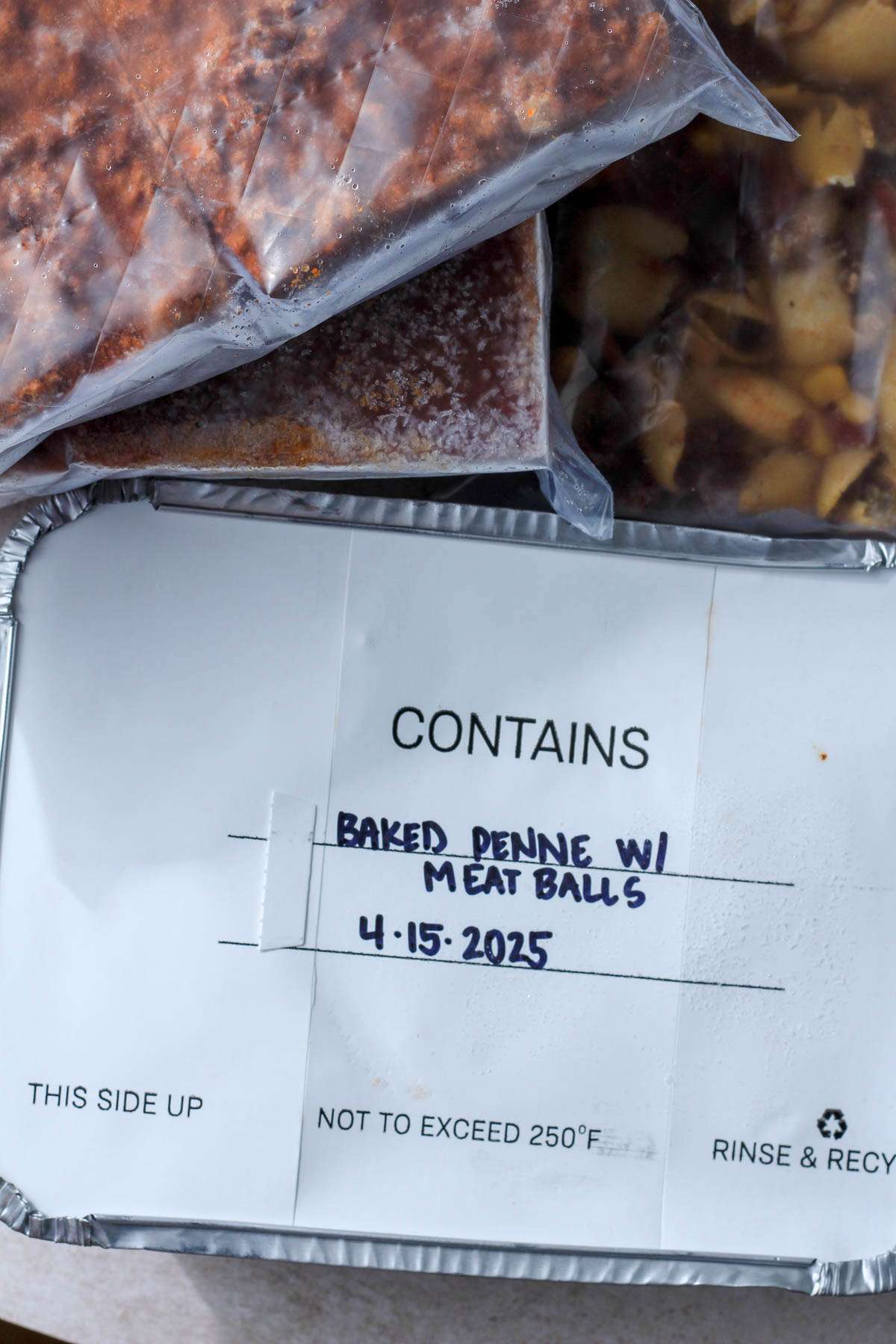 A small tray of baked penne with meatballs and bags of other freezer foods around it.