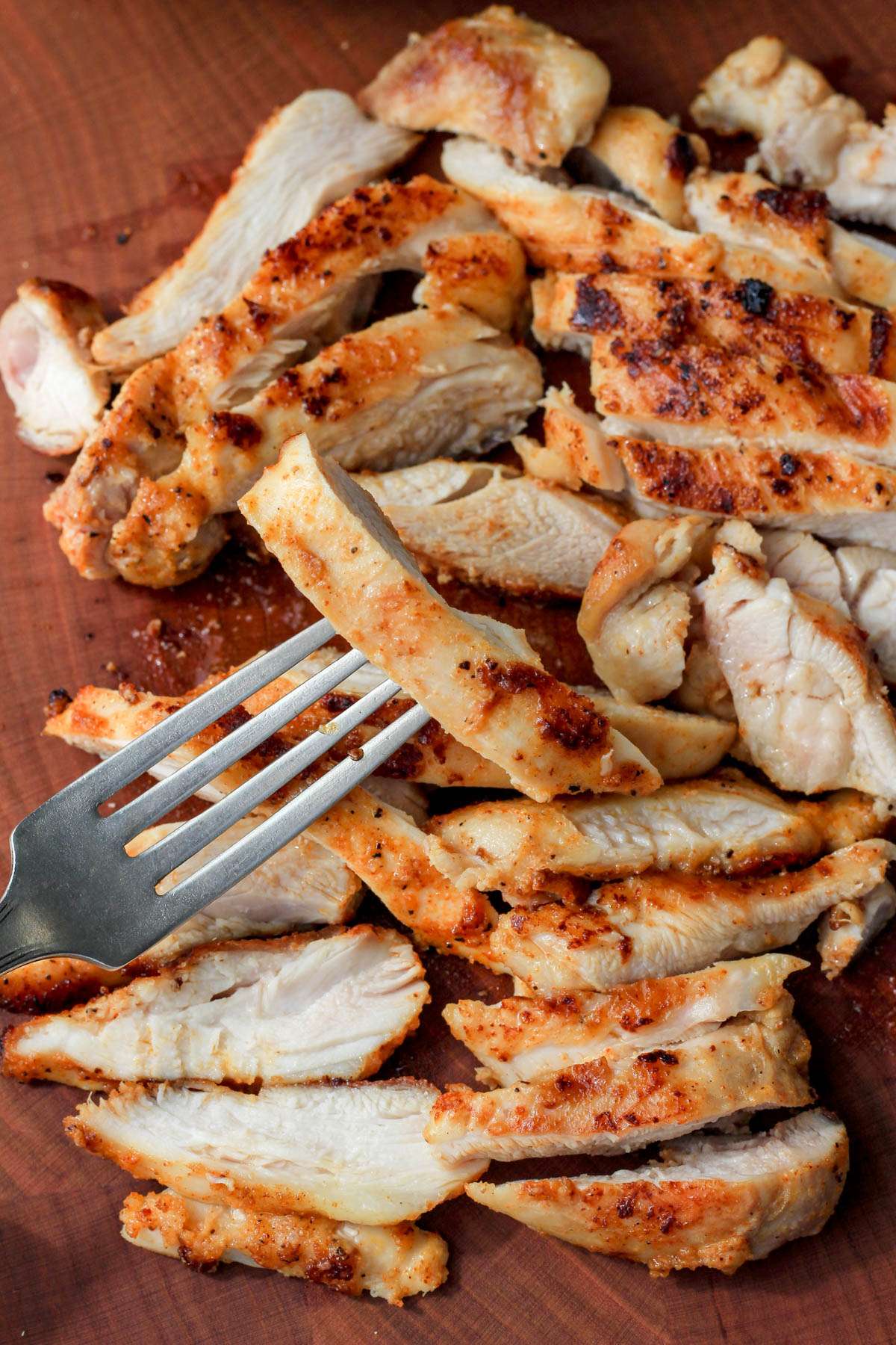 A silver fork with a boneless skinless chicken thigh piece in front of a pile of chicken on a wooden cutting board.