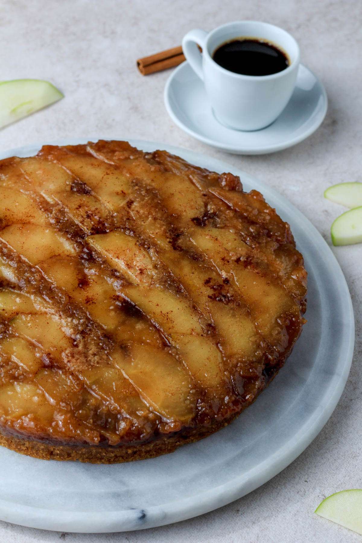 A marble cake stand with an apple upside down cake and a small white cup of coffee in the back right.
