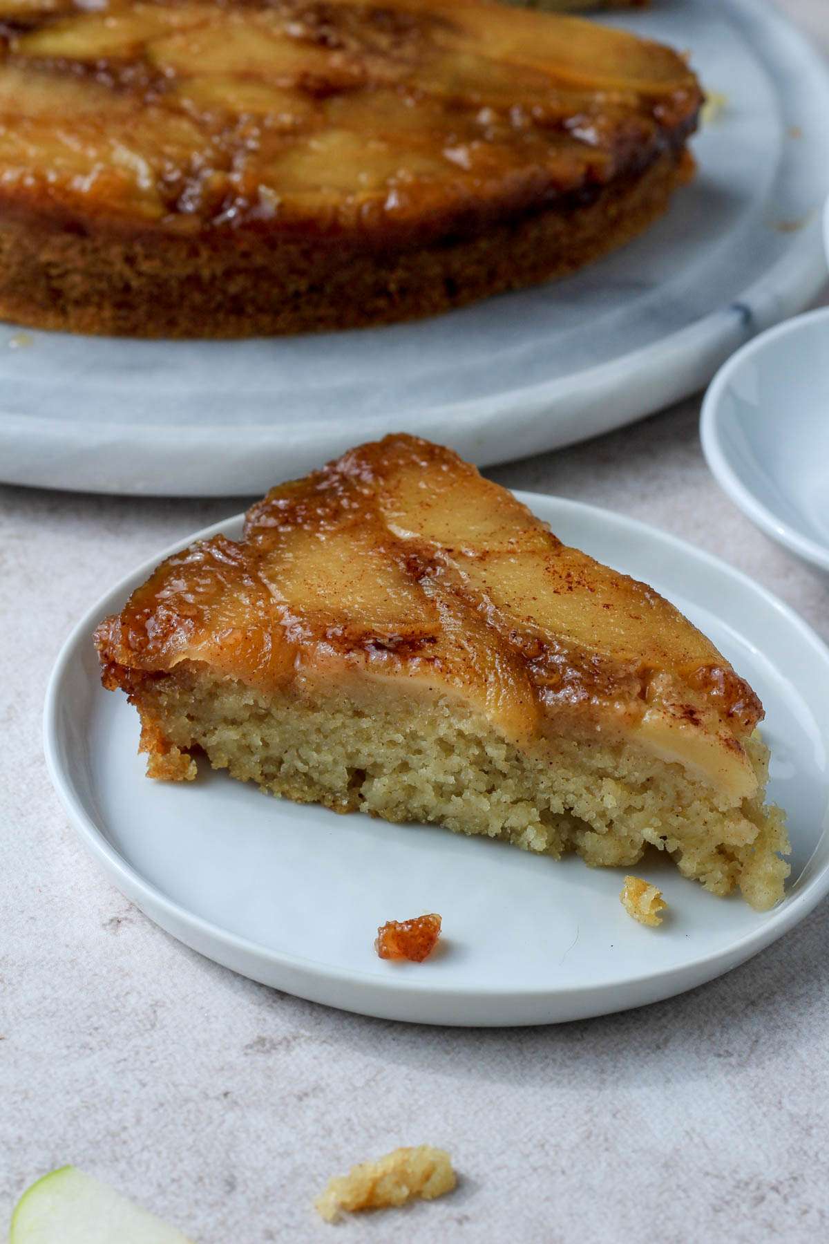 A small white plate with a slice of vegan cinnamon apple upside down cake in front of a marble cake stand with the remaining cake.