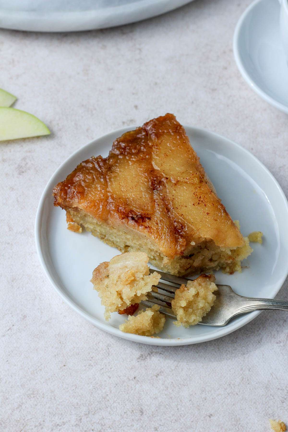 A small white plate with a slice of vegan cinnamon apple upside down cake with a bite taken out on a silver fork to the left of the slice.