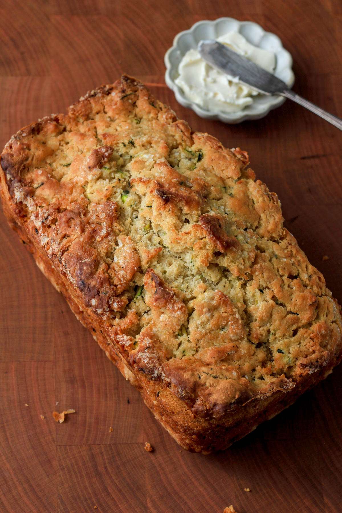 A wooden cutting board with a loaf of zucchini beer bread next to a small dish of vegan butter with a small silver butter knife to the top right.
