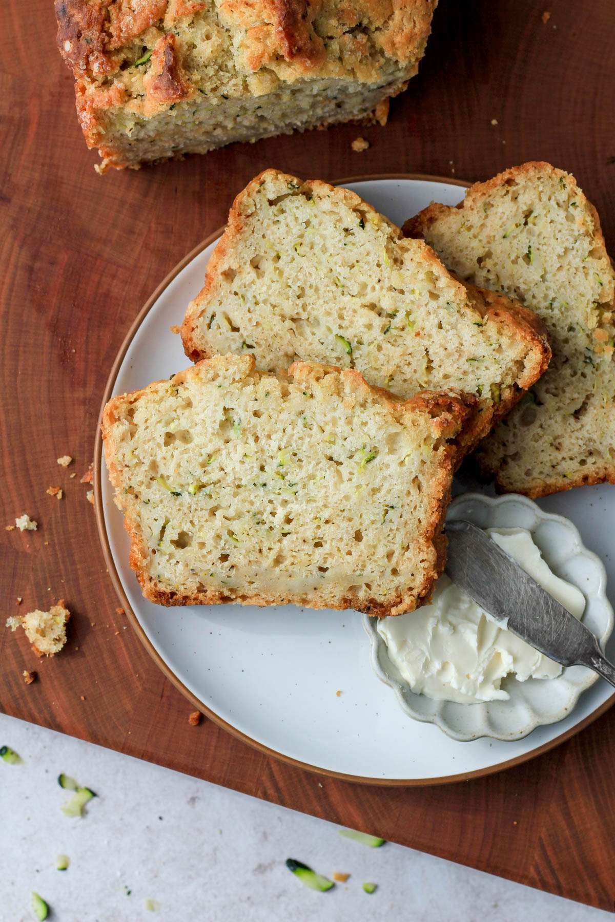 A white plate on top of a wooden cutting board with three slices of zucchini beer bread and a small dish of vegan butter with a silver butter knife.
