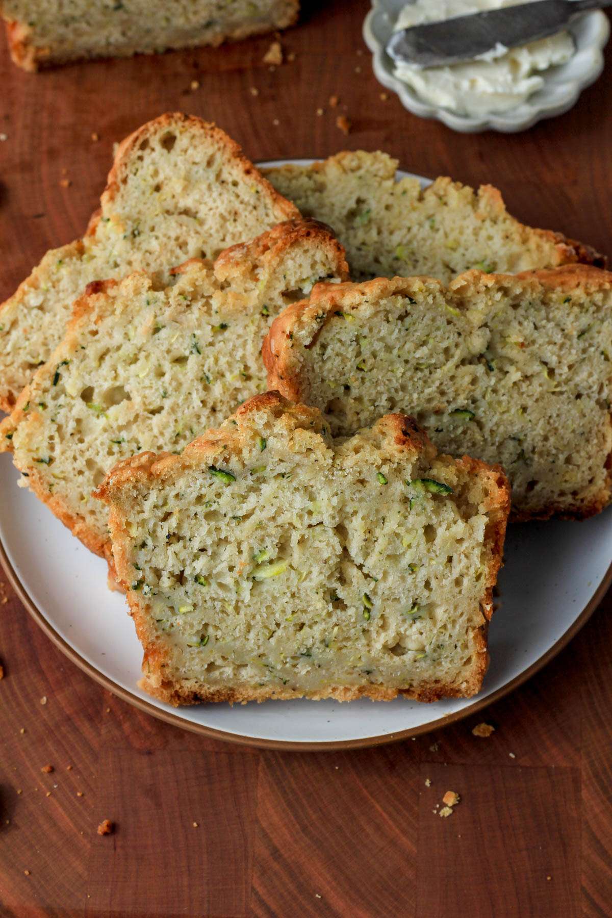 A wooden cutting board with a white plate on top with five slices of zucchini beer bread and a small dish of vegan butter in the back right.