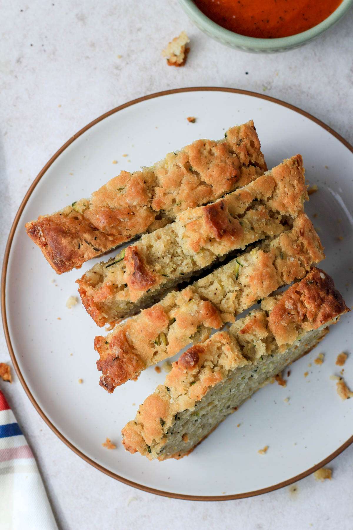 A white plate with a brown rim and four slices of zucchini beer bread with a small bowl of tomato soup in the top right corner.