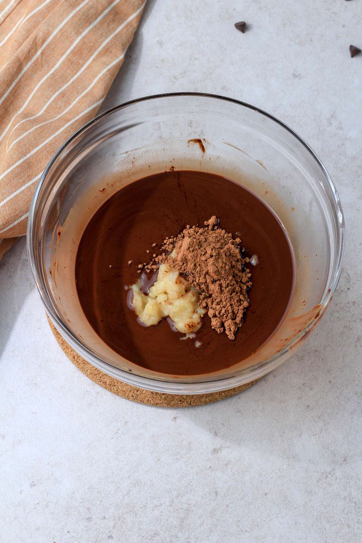 A glass mixing bowl with melted chocolate topped with cocoa powder and a mashed banana.