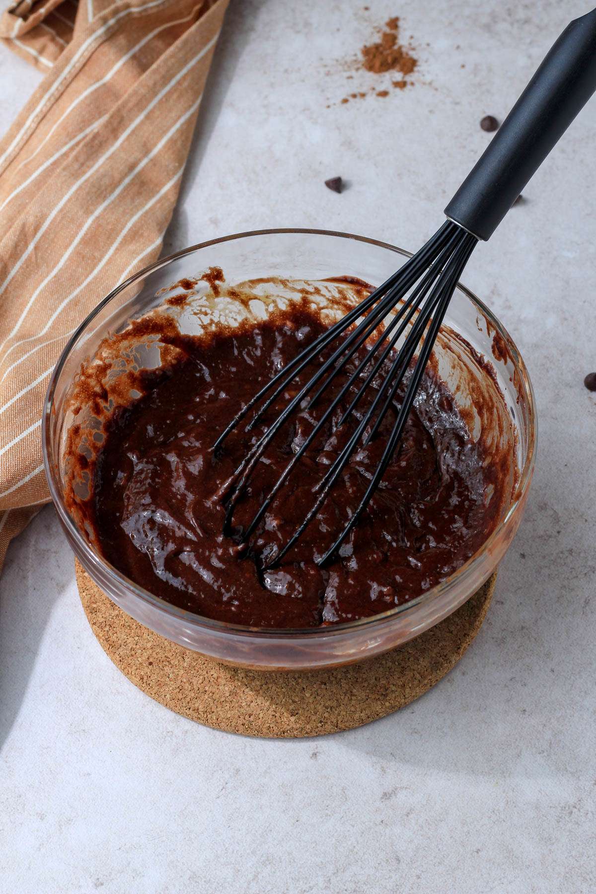 Chocolate brownie batter in a glass mixing bowl with a black whisk on a tan counter.