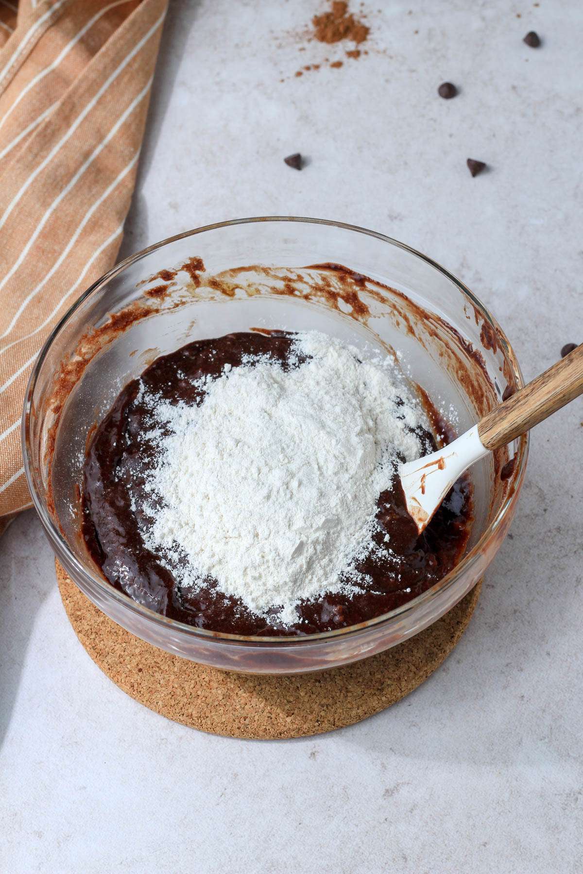 A glass mixing bowl with brownie batter topped with flour before folding in.
