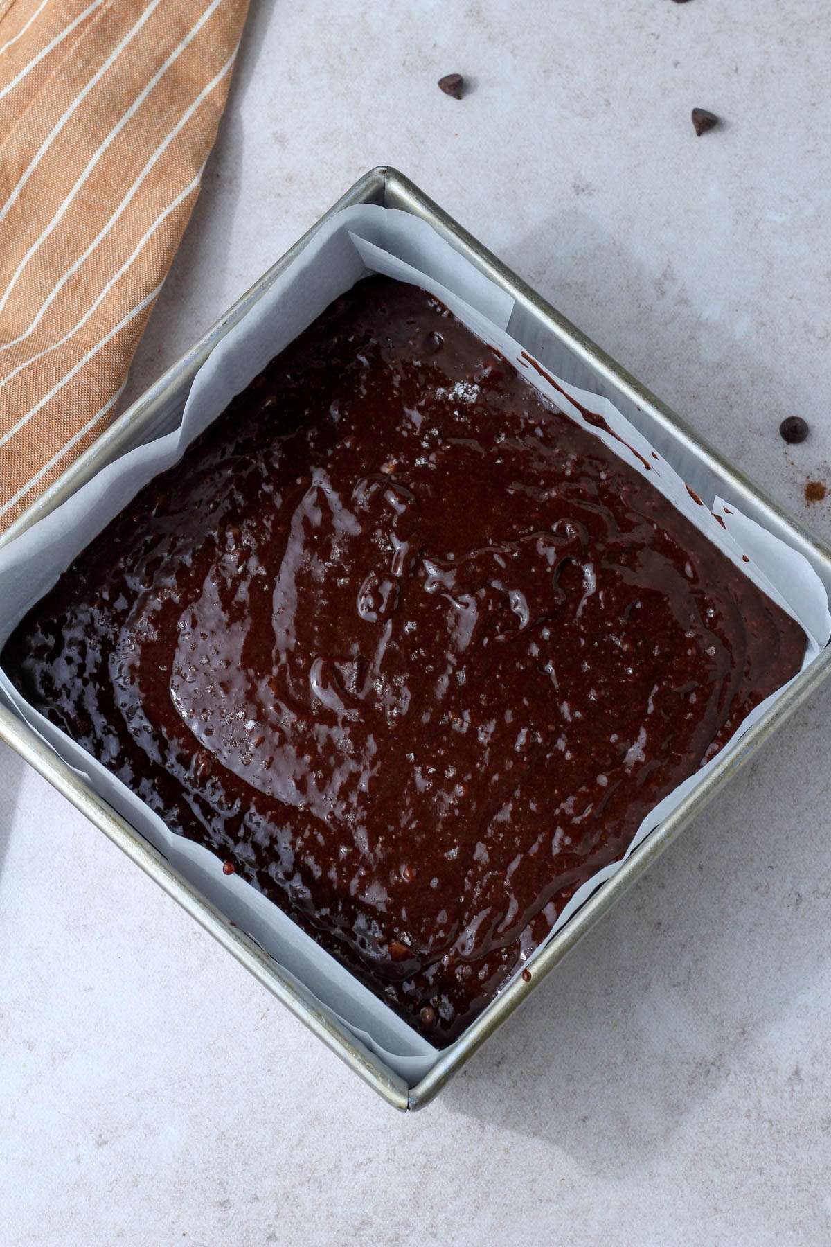 A silver square baking pan lined with parchment paper and filled with dairy-free brownie batter.