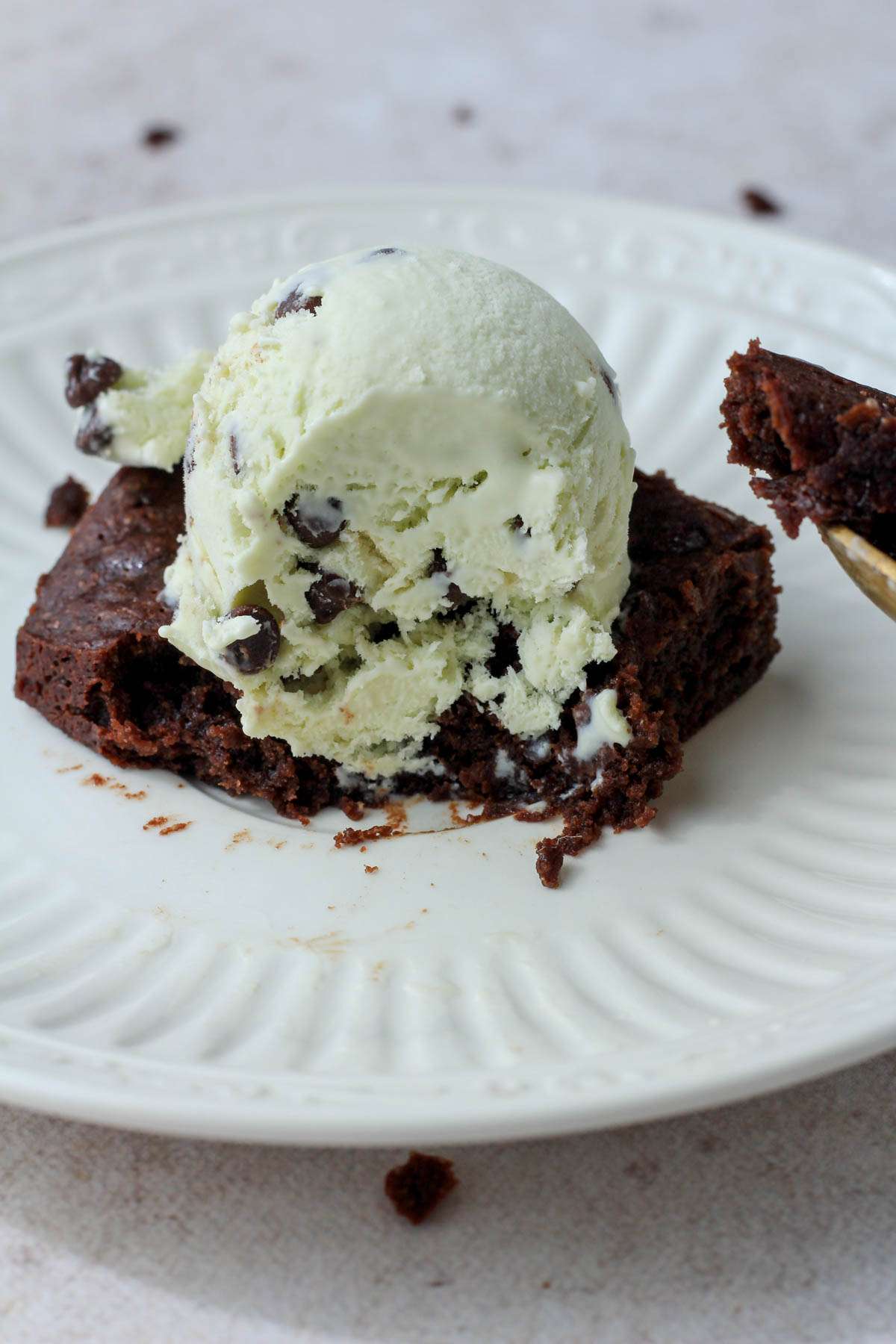 A white plate on a cream counter with a dairy-free chocolate brownie topped with a scoop of mint chocolate chip ice cream.