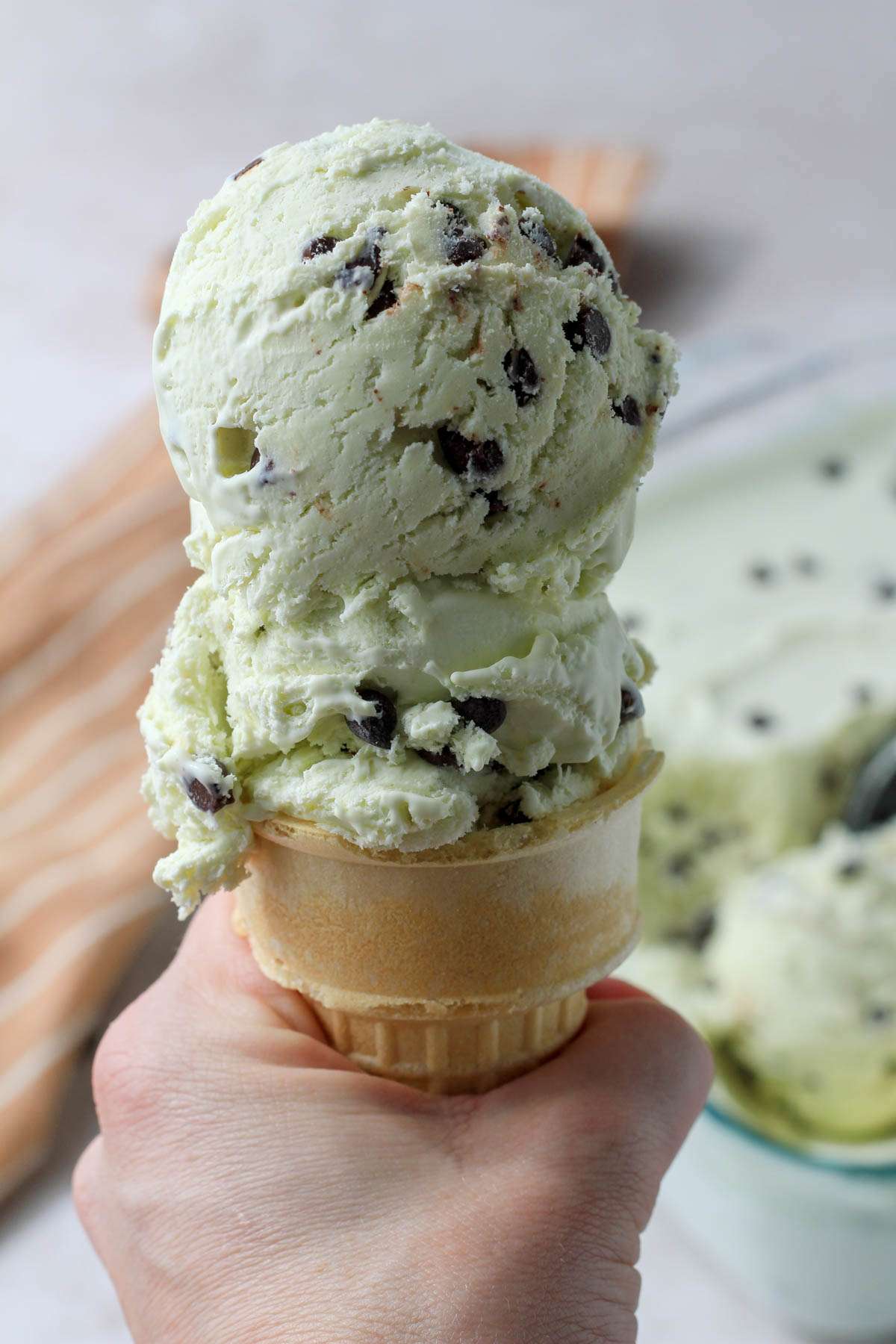 A hand holding a cake cone filled with two scoops of no churn mint chocolate chip ice cream.
