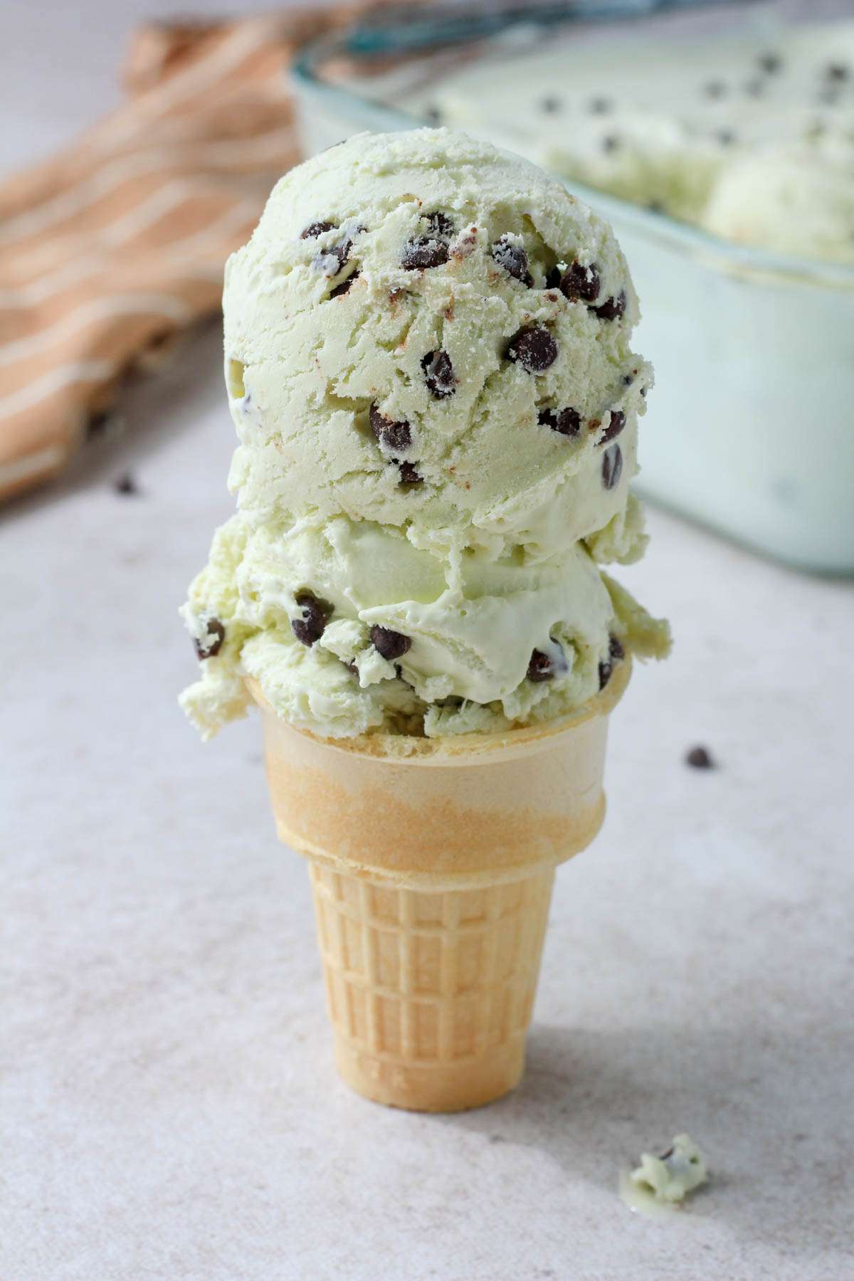 A cake cone on a tan counter with two scoops of mint chocolate chip ice cream on the cone.