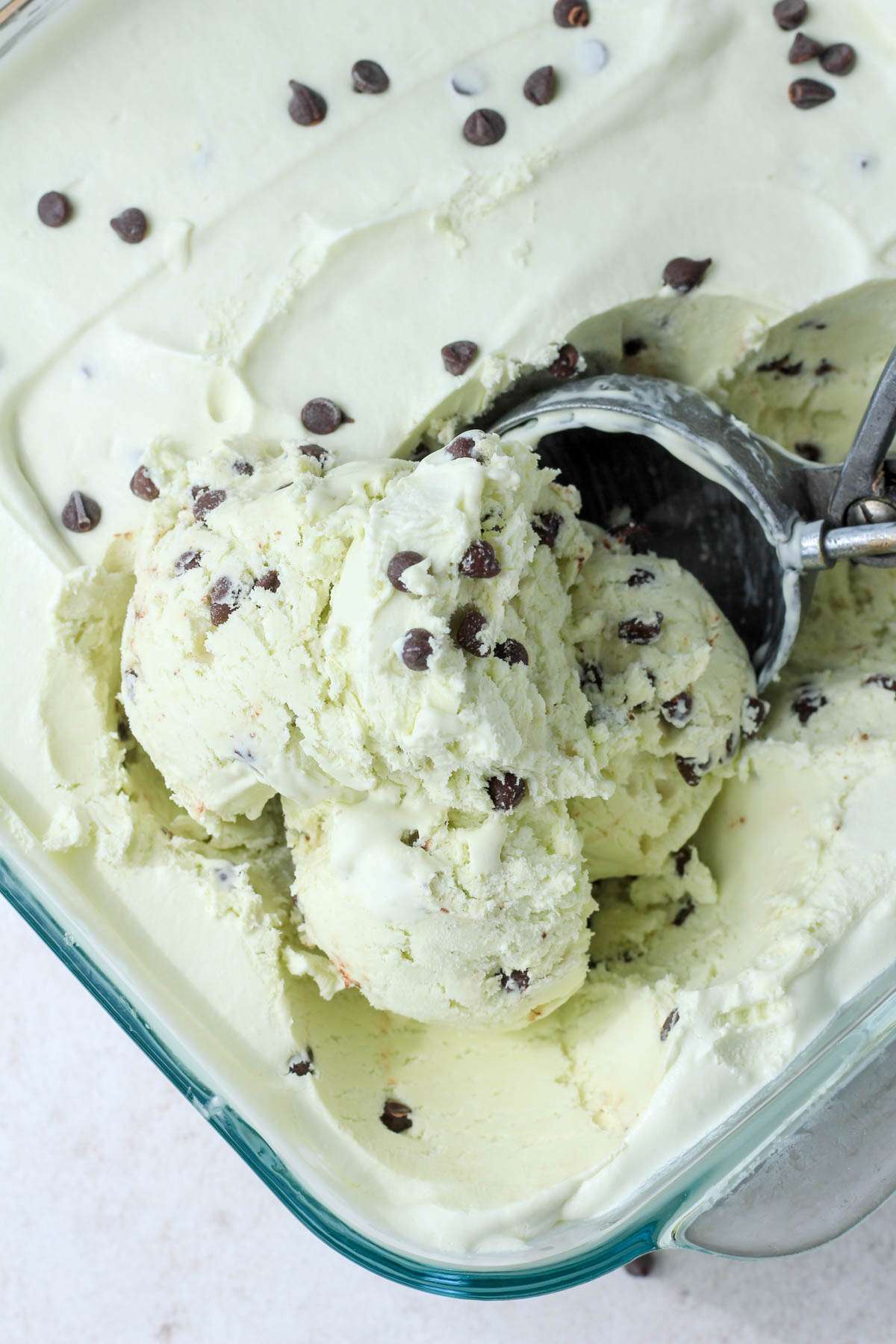A glass dish with vegan mint chocolate chip ice cream and a silver ice cream scoop scooping balls of ice cream.