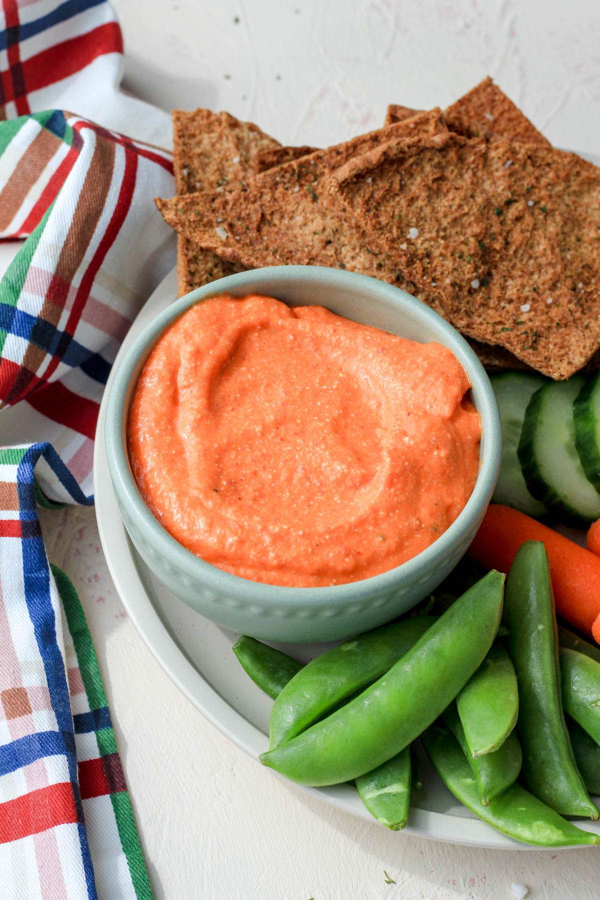 A small green bowl filled with vegan feta and red pepper dip on a plate with snap peas, pita chips, and carrots.