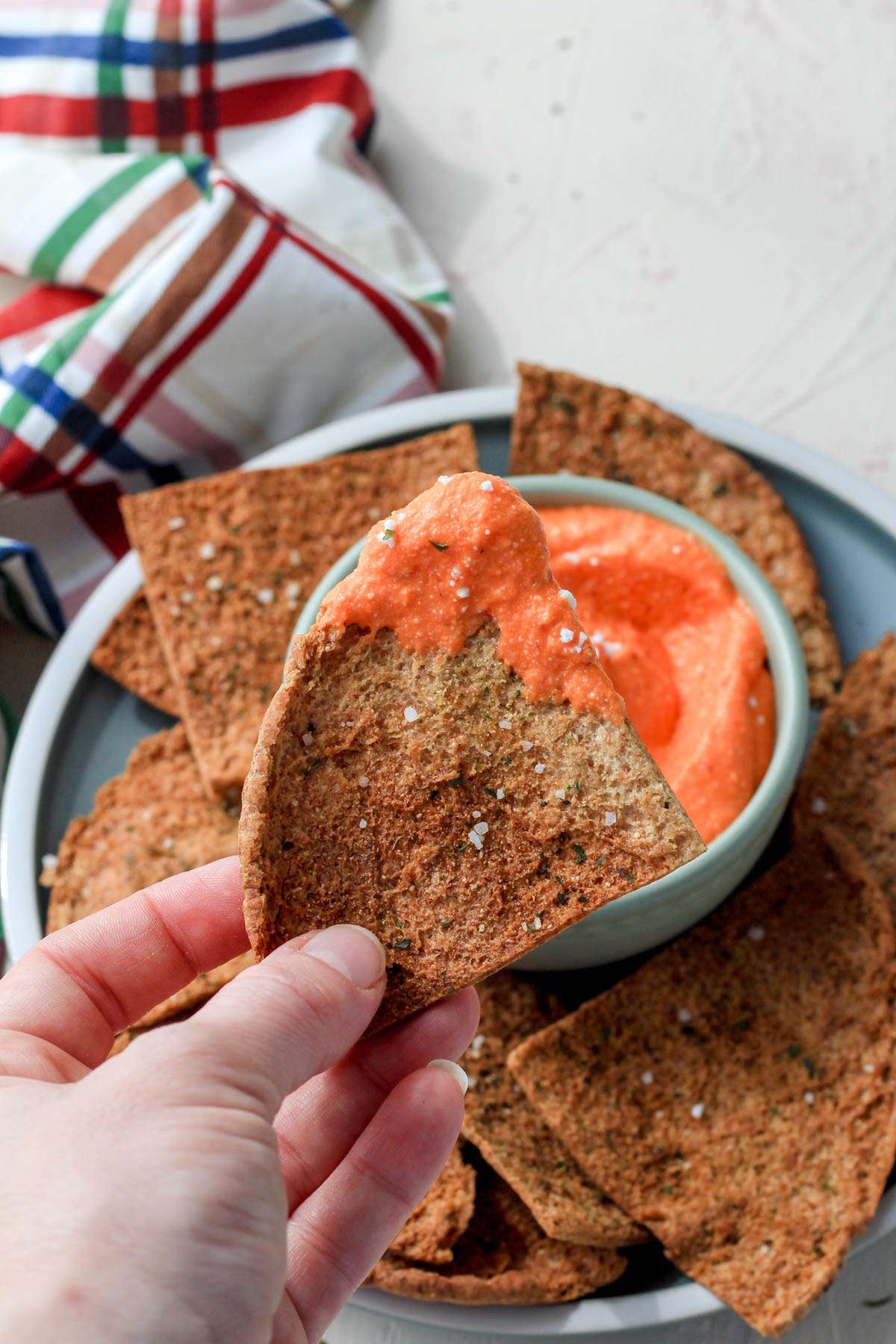 A hand holding a pita chip with a scoop of vegan roasted red pepper and feta dip over a bowl of dip.