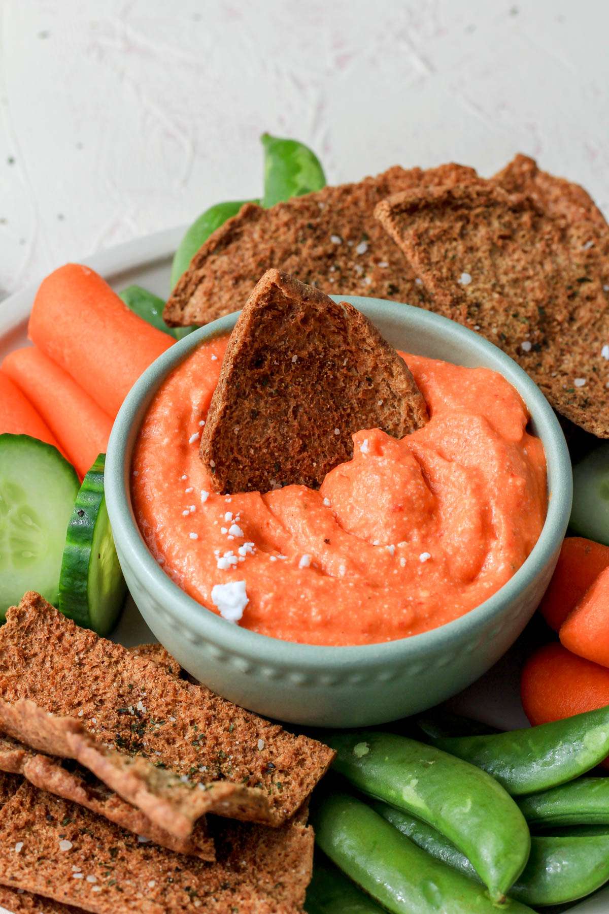 A crispy pita chip dipped into a small green bowl with vegan roasted red pepper dip with sprinkles of feta on a plate surrounded by vegetables.