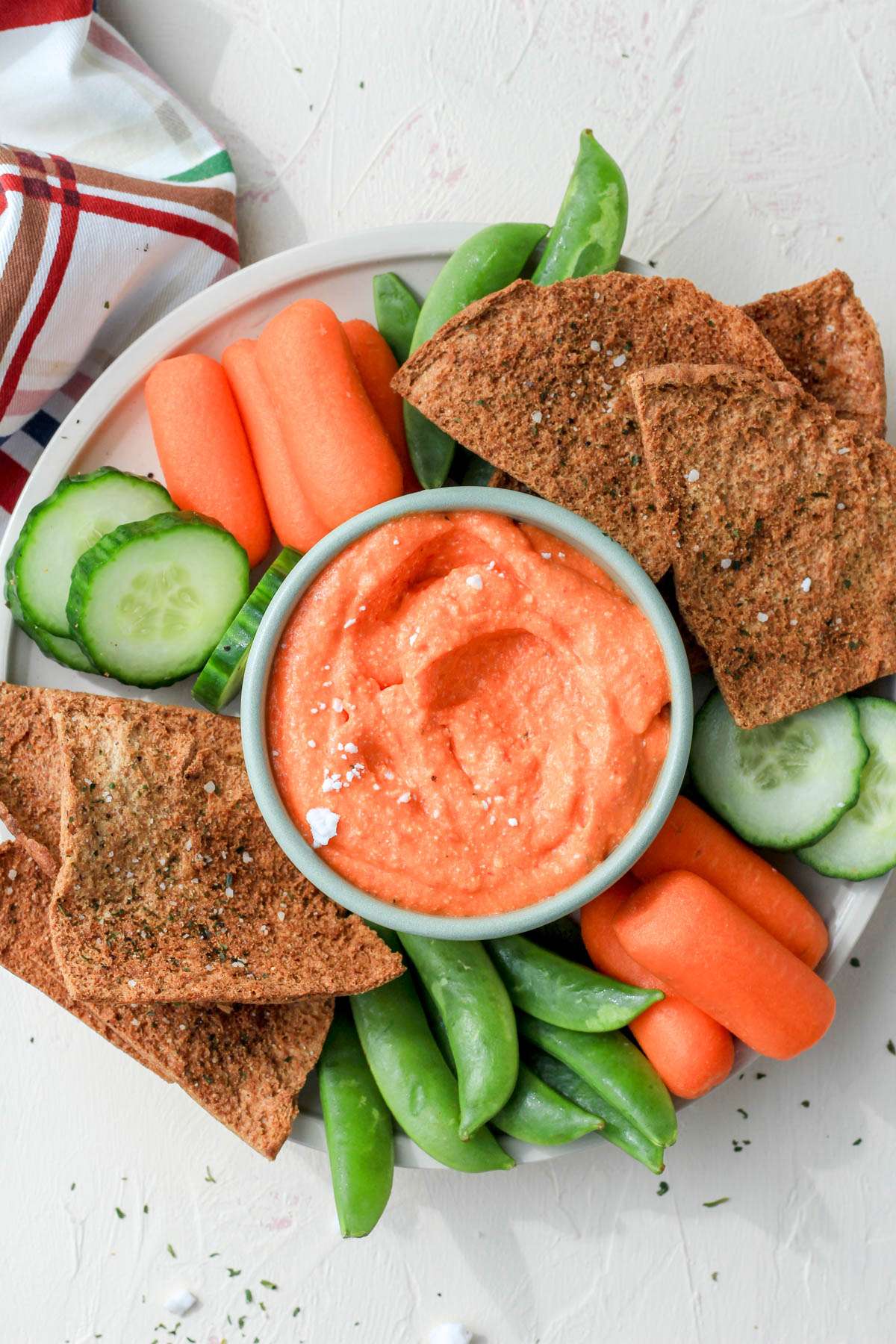 A top down image of a small green bowl of vegan feta and roasted red pepper dip surrounded by vegetables and pita chips.