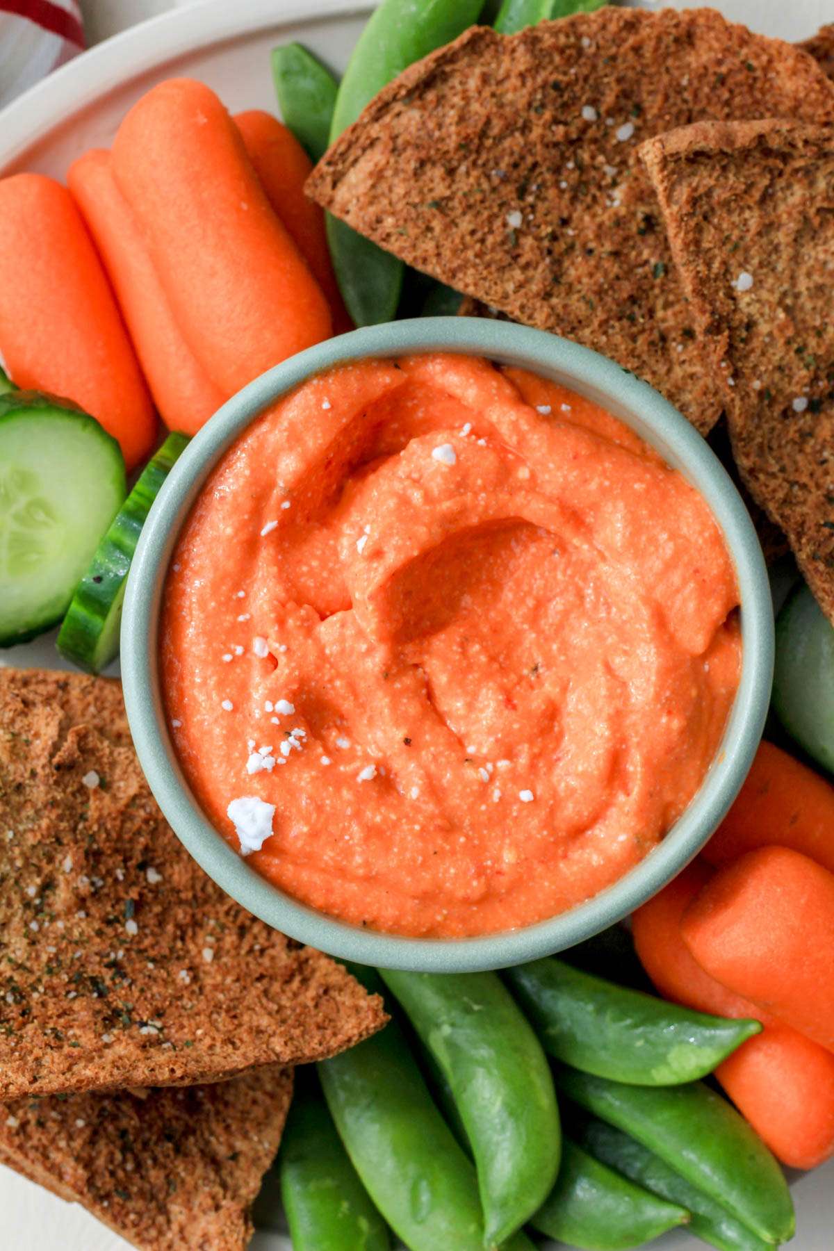 A close up of a green bowl with creamy red pepper dip topped with some vegan feta with snap peas, carrots, and pita chips surrounding the bowl.
