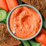 A light green bowl filled with vegan roasted red pepper dip surrounded by vegetables and pita chips.