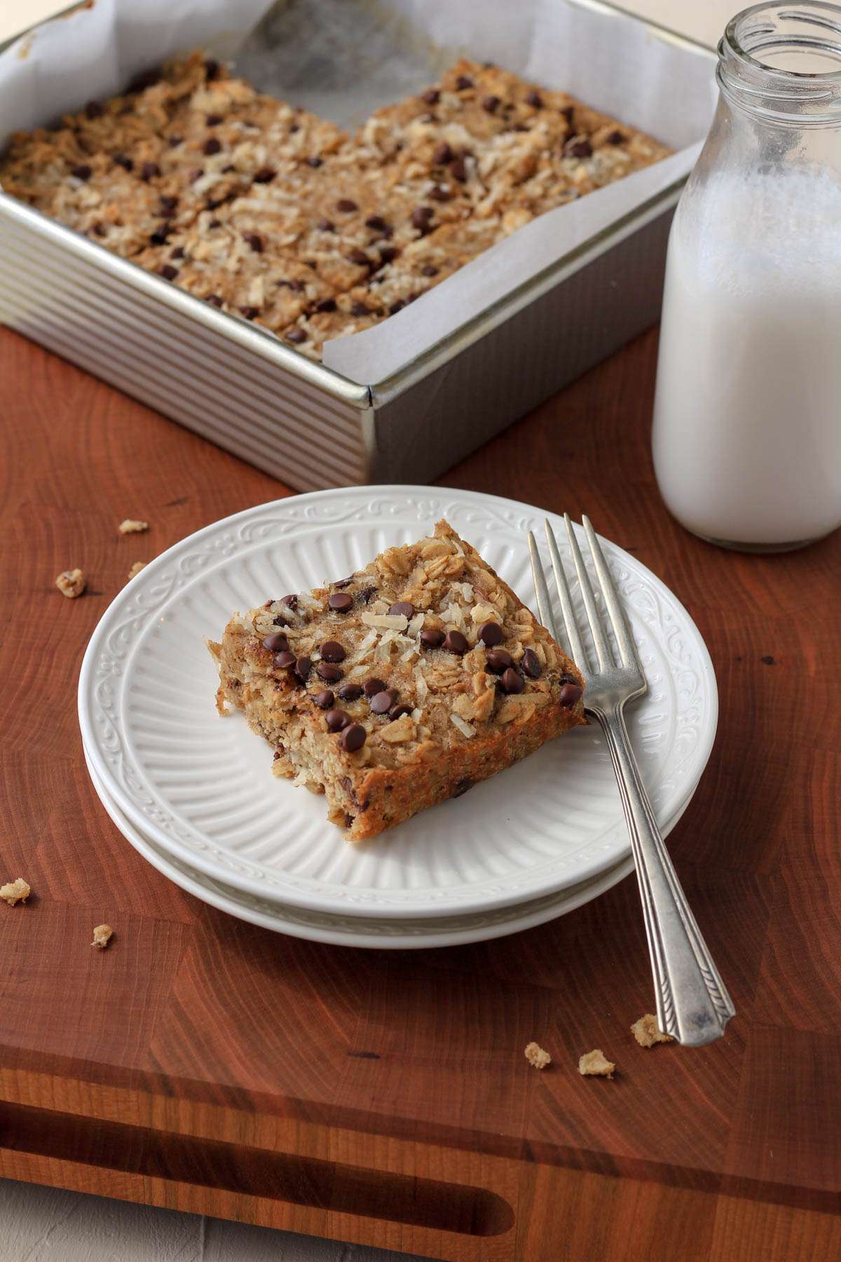 A wooden cutting board topped with a white plate of almond joy baked oatmeal with a silver fork and a dish of baked oatmeal in the back left with a jut of almond milk in the back right.