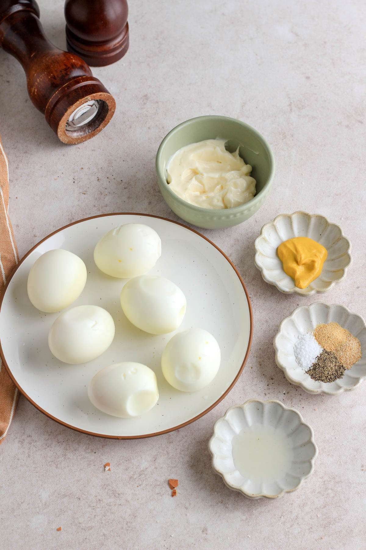 A white plate with six hard boiled eggs next to bowls with the ingredients for making deviled eggs.