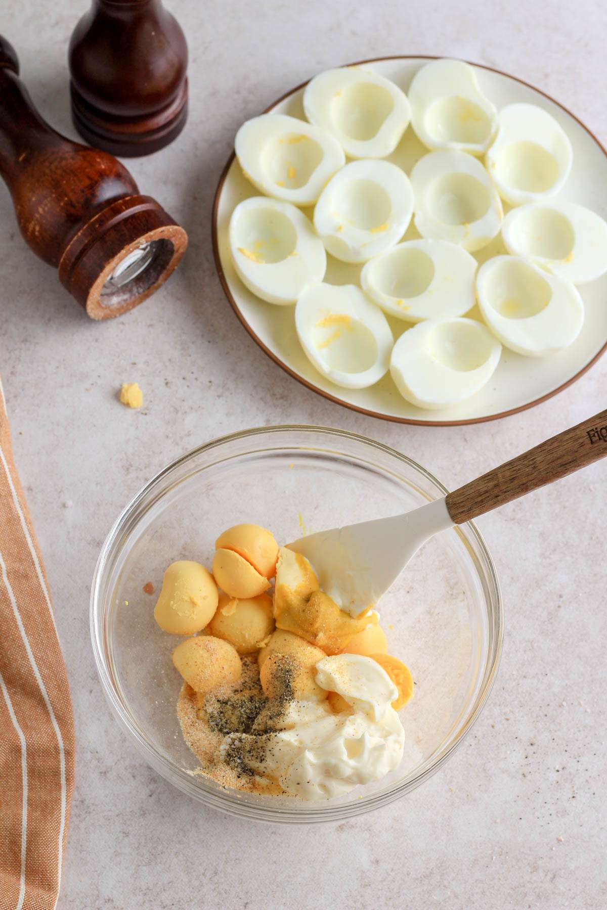 A small glass bowl with cooked eggs yolks, mayo, mustard, and spices for deviled egg filling with a plate of egg white halves in the back right.