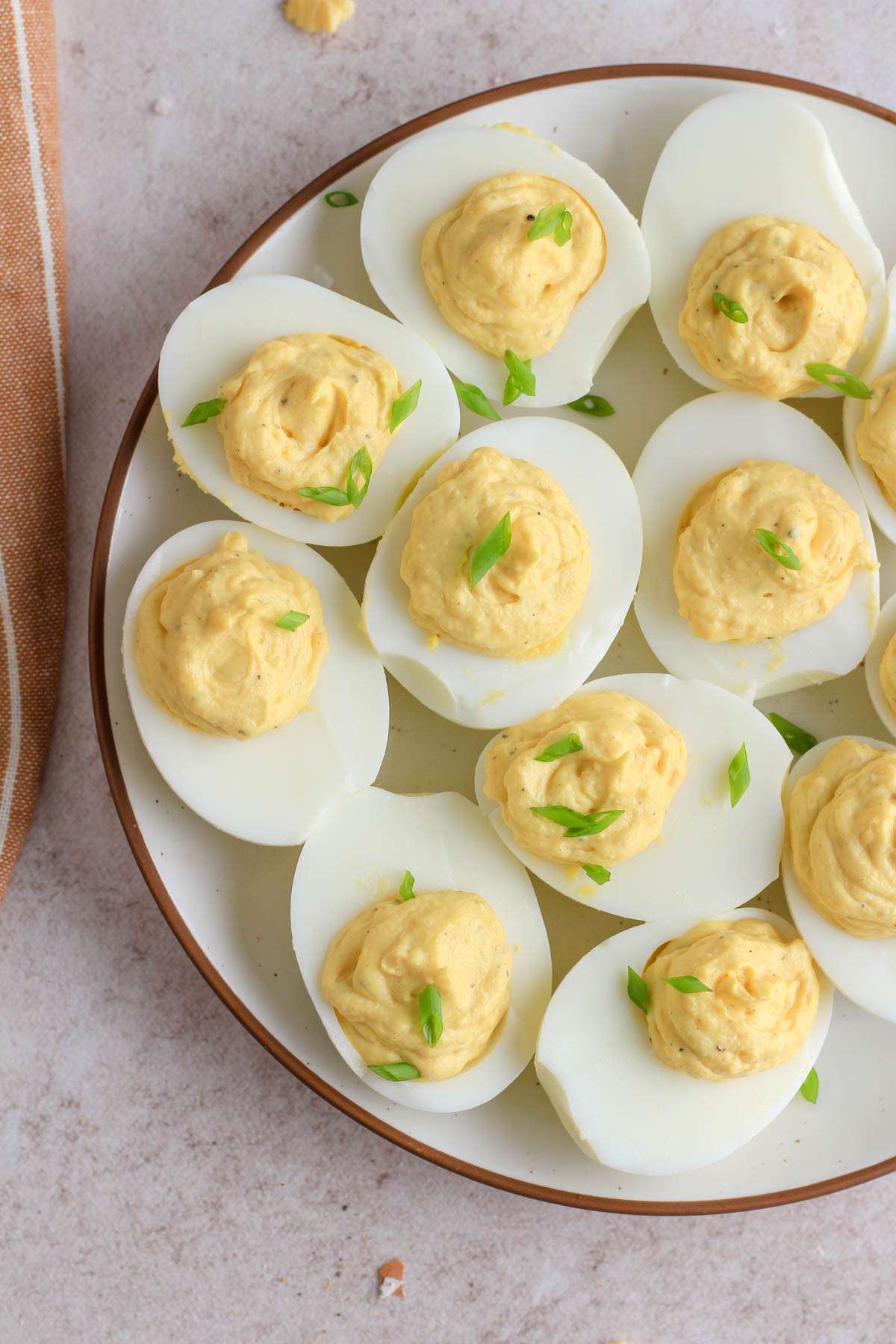 Classic deviled eggs topped with sliced green onion on a white plate with a brown rim on a tan counter.