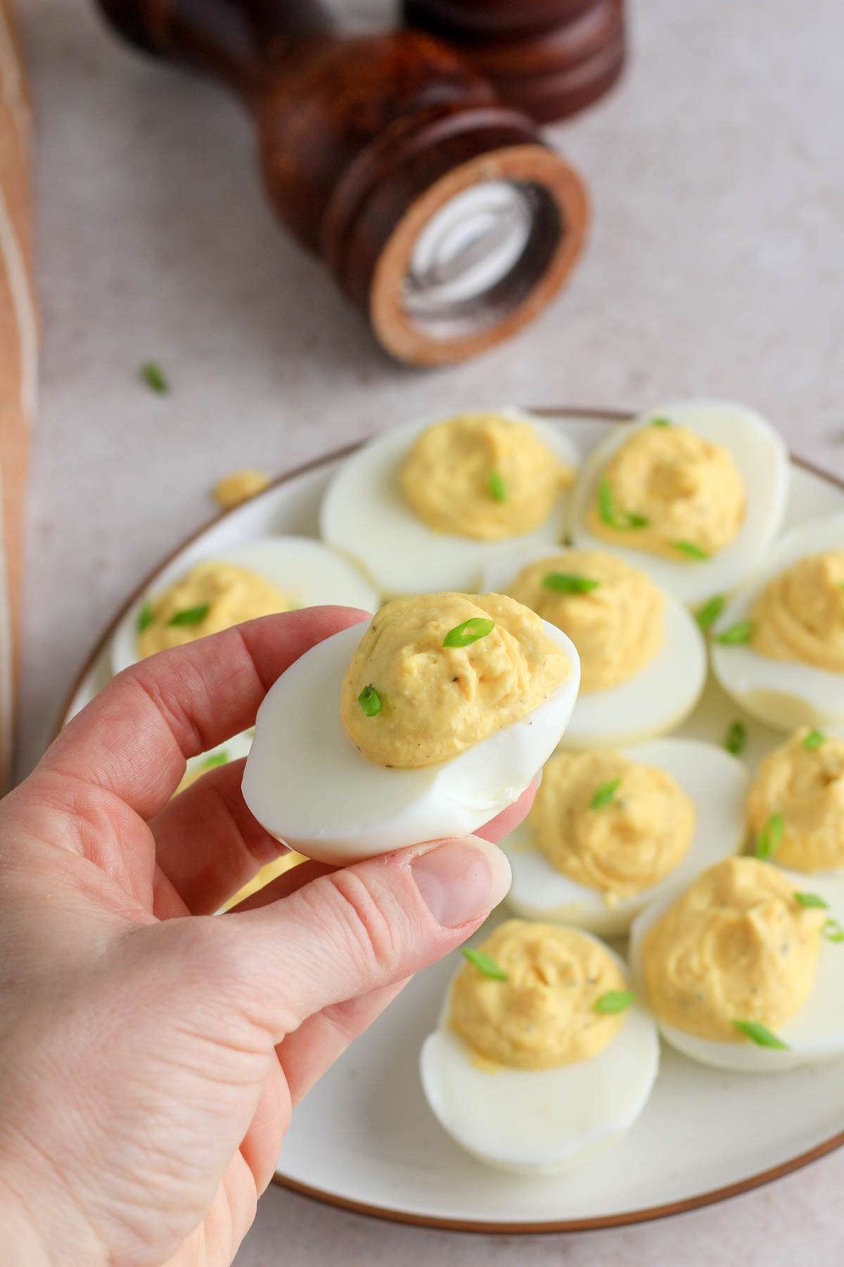 A hand holding a classic deviled egg topped with sliced green onion over a platter of other deviled eggs.