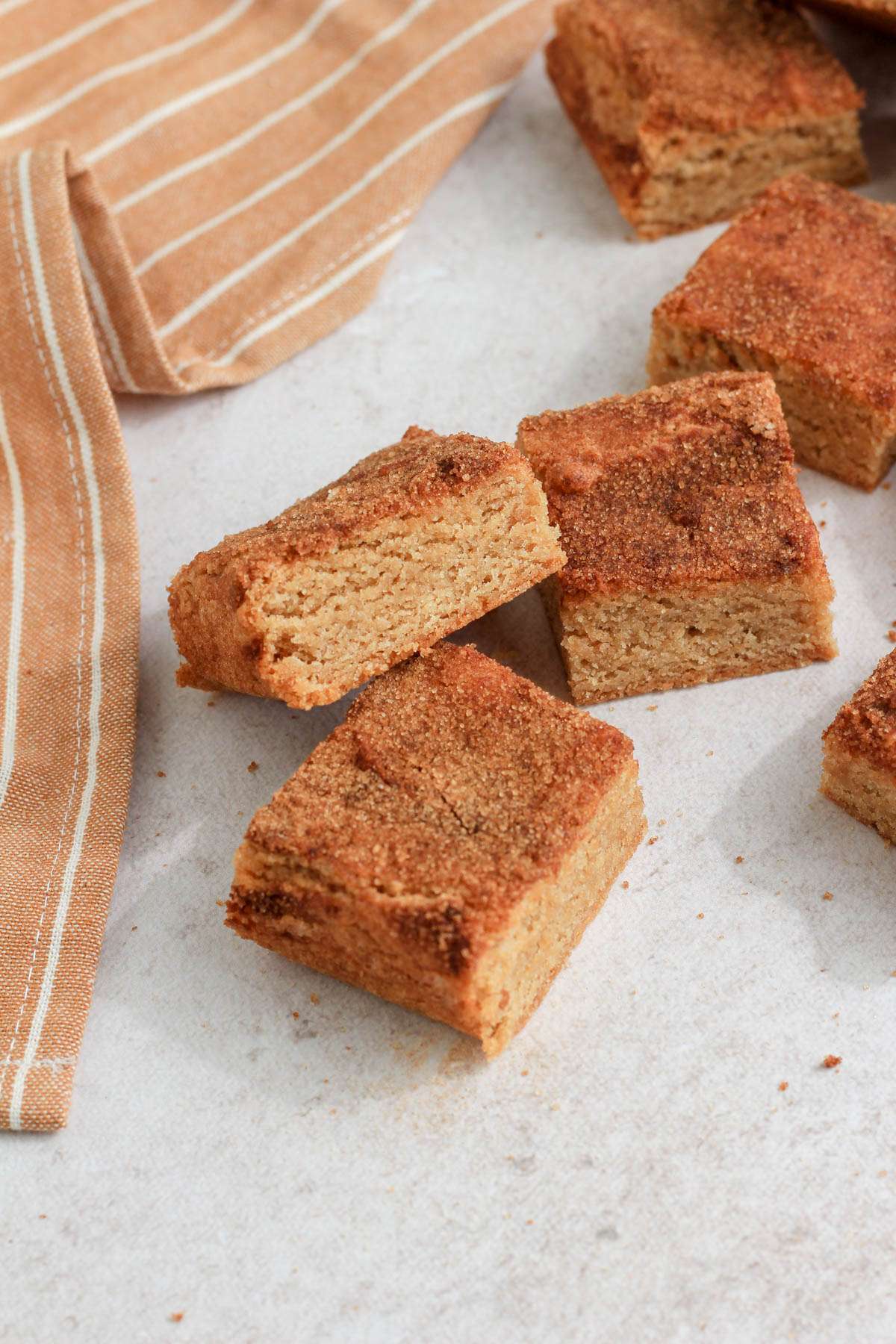 A small pile of snickerdoodle cookie bars on a tan counter with cookie crumbs on the counter and an orange and white striped towel on the left side.