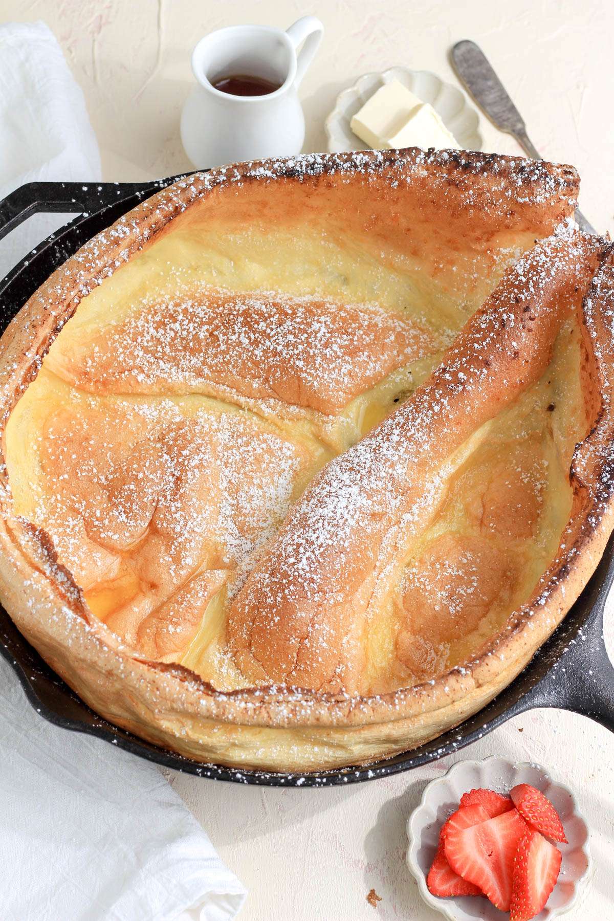 A black cast iron skillet on a cream and pink counter with a dutch baby dusted in powdered sugar with a small jug of maple syrup in the back left and a dish of sliced strawberries in the front right.