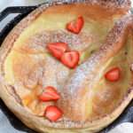 A close up of a dairy-free dutch baby in a skillet with powdered sugar and sliced strawberries.