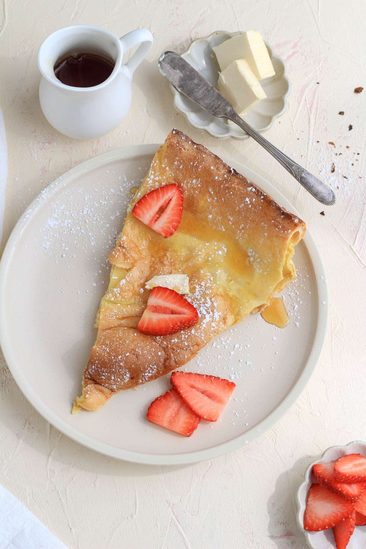 A cream plate with a slice of a dutch baby topped with powdered sugar and strawberries.