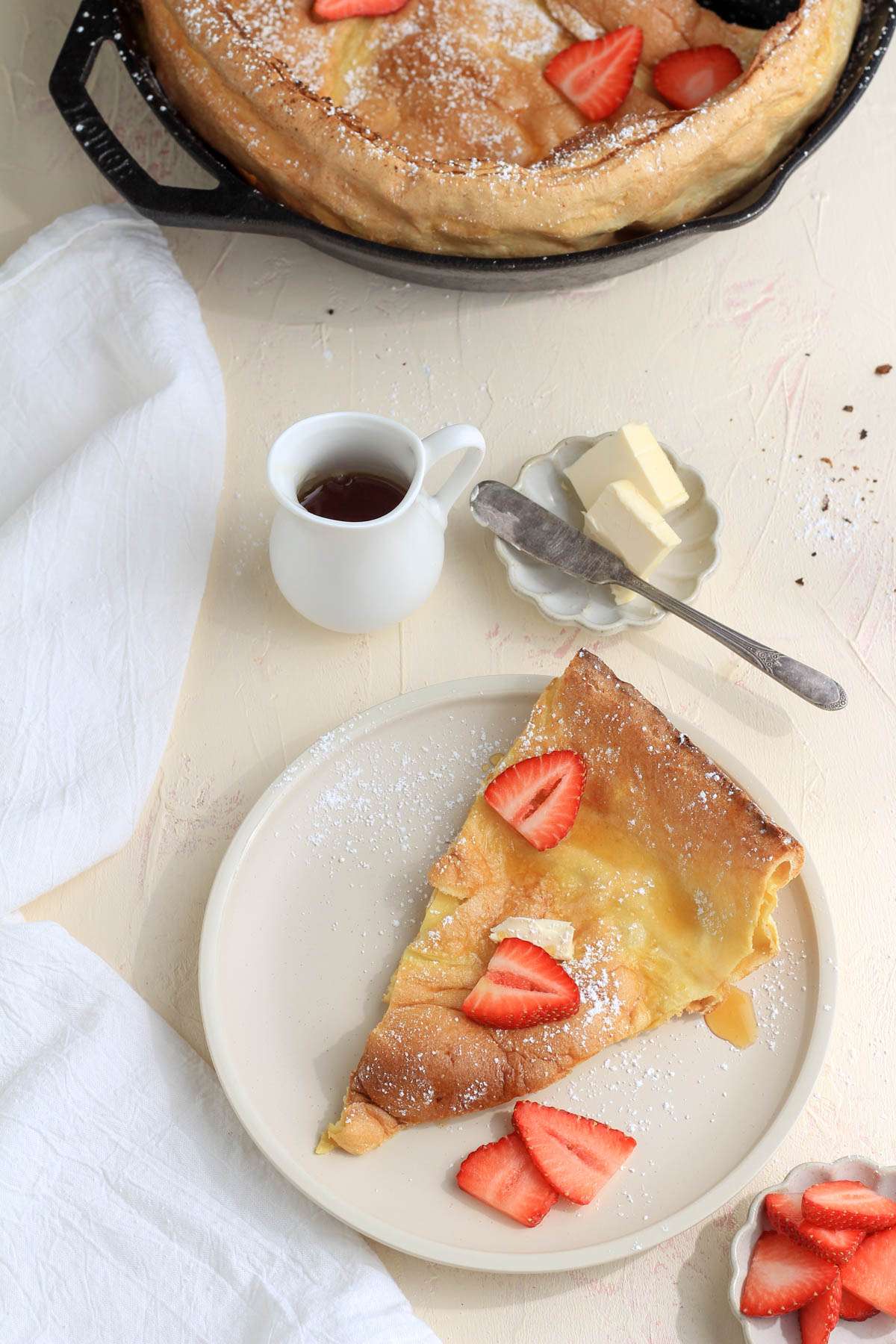 A counter with a bowl of strawberries, a plate with a wedge of dutch baby, a small jug of maple syrup, and a cast iron skillet with the remaining dutch baby.