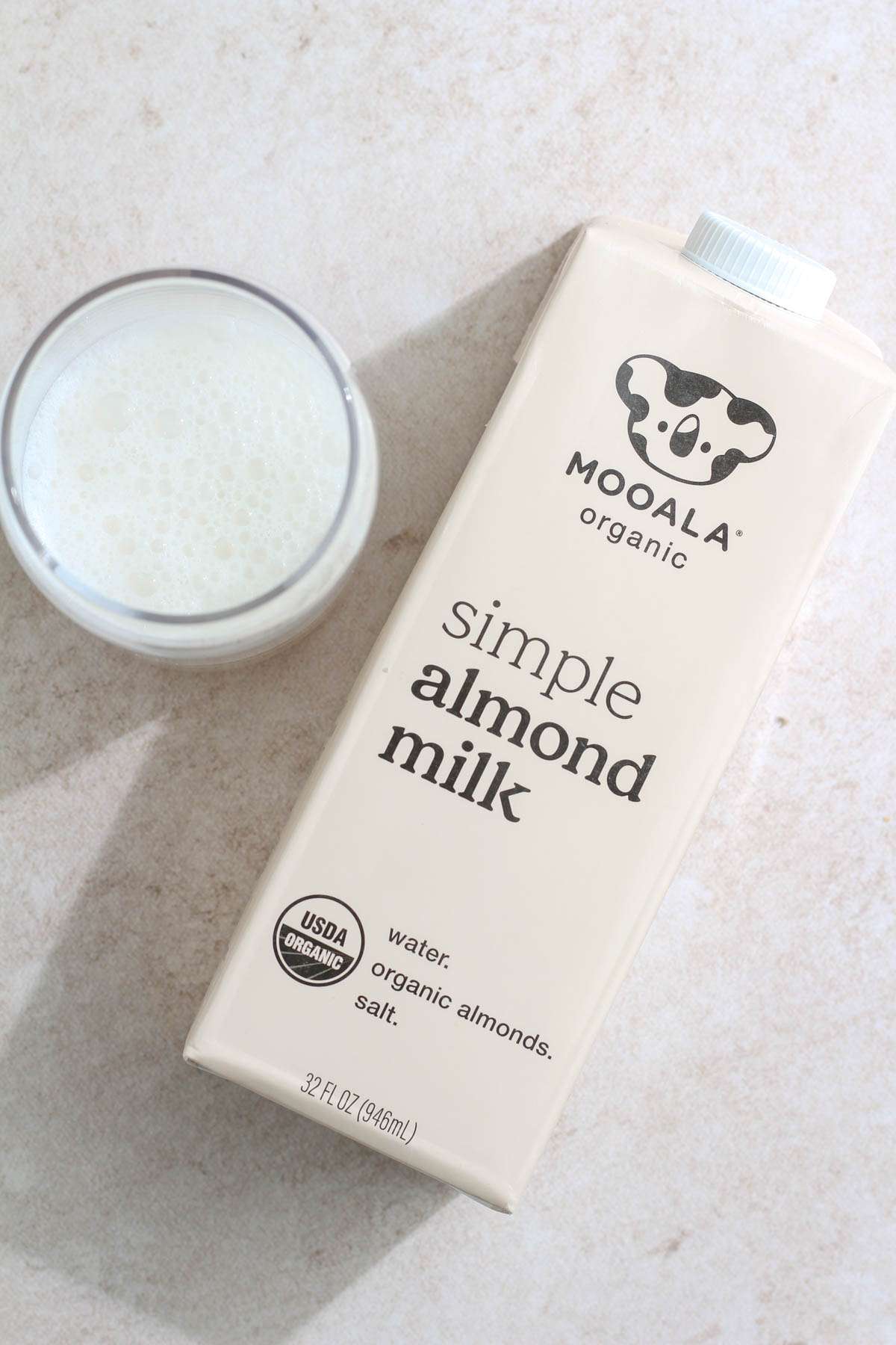 A glass cup with a serving of Mooala Almond milk next to a tan carton of Mooala almond milk.