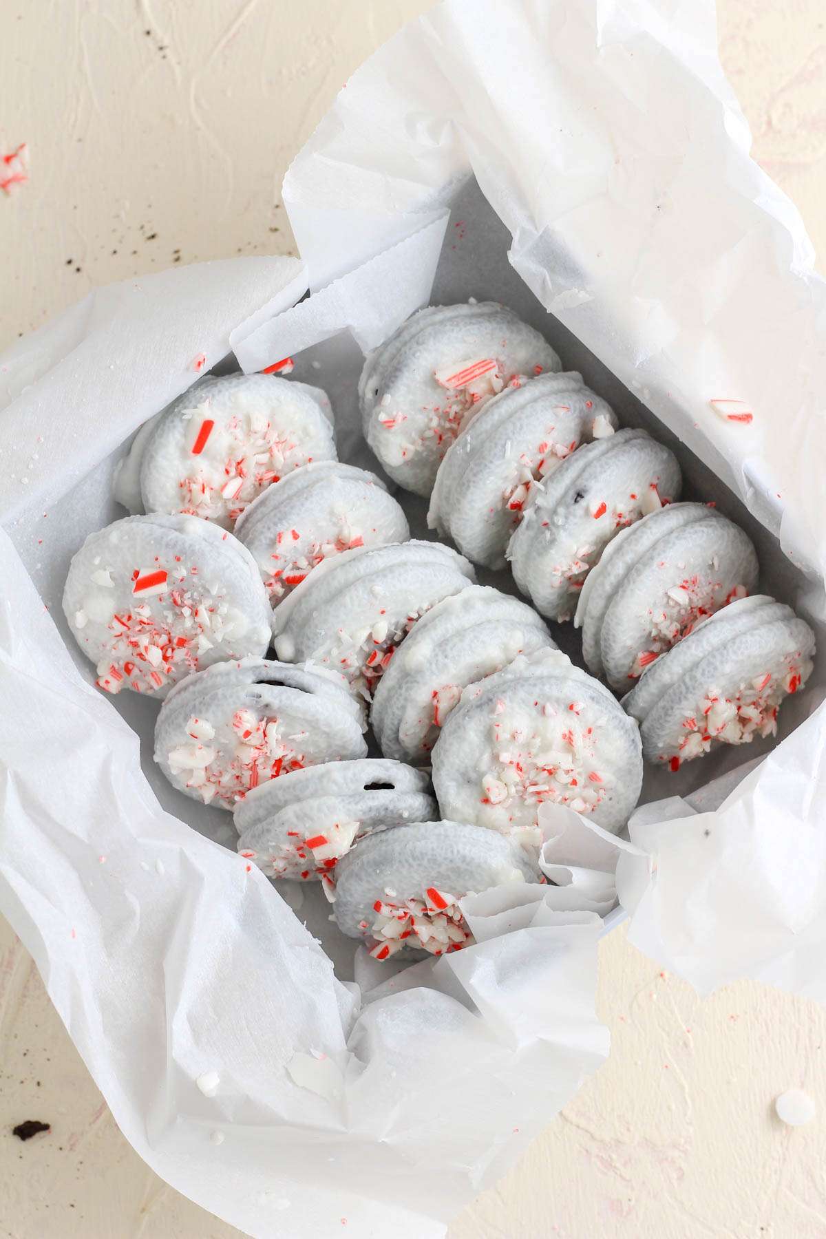 Rows of vegan peppermint chocolate oreos topped with crushed candy cane in a parchment paper lined cookie box on a cream counter.