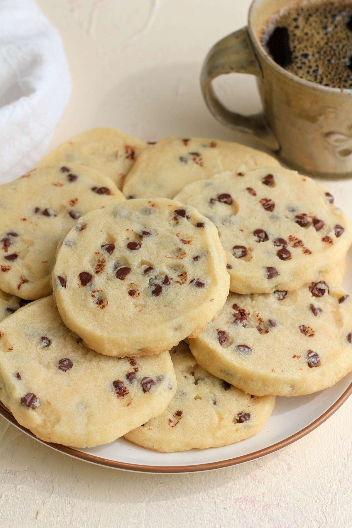 A cream counter with a white plate topped with vegan chocolate chip cookies and a small cup of coffee in the back right.