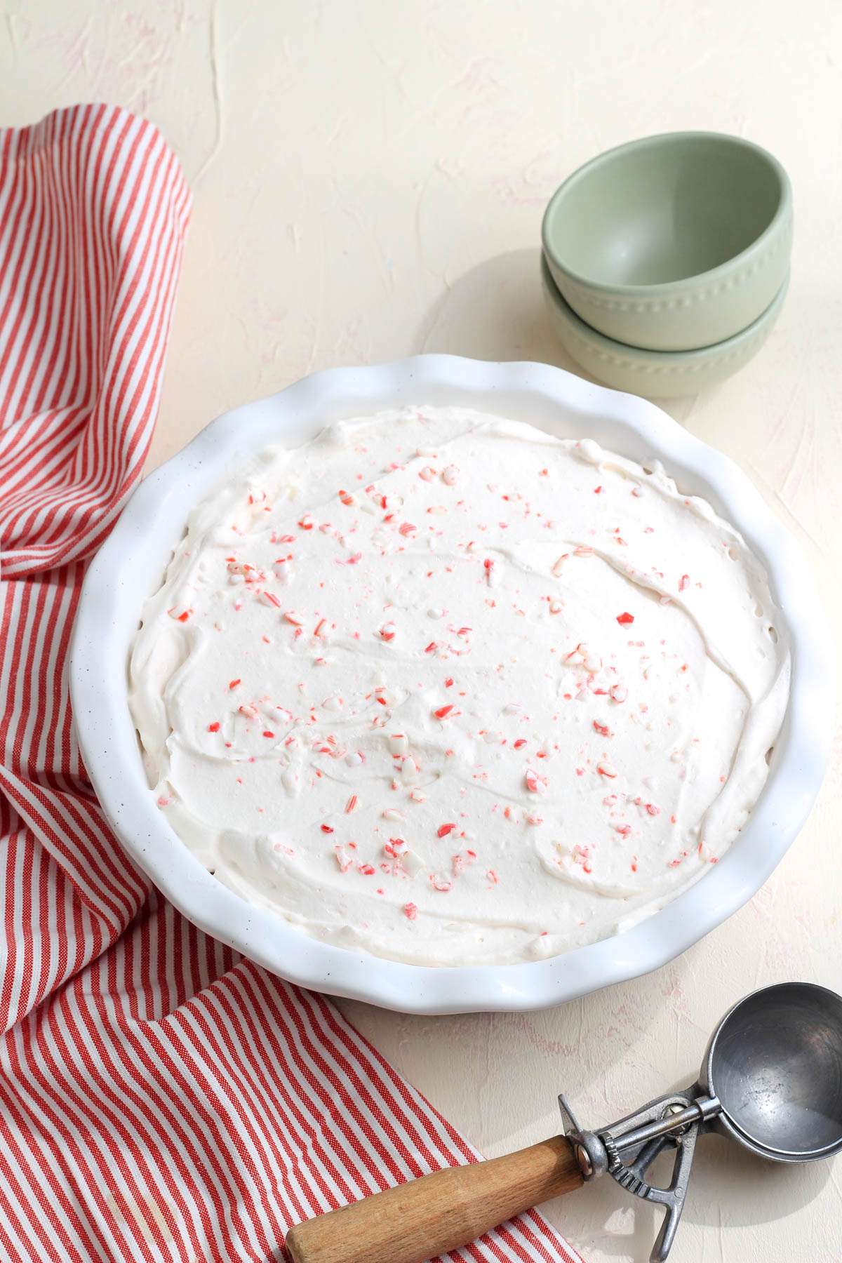 A deep white pie pan on a cream counter with an ice cream scoop and a stack of green bowls in the back right with frozen no churn peppermint ice cream.