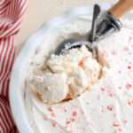 A close up of a white pan filled with vegan peppermint ice cream and a silver and wooden ice cream scoop scooping the ice cream.