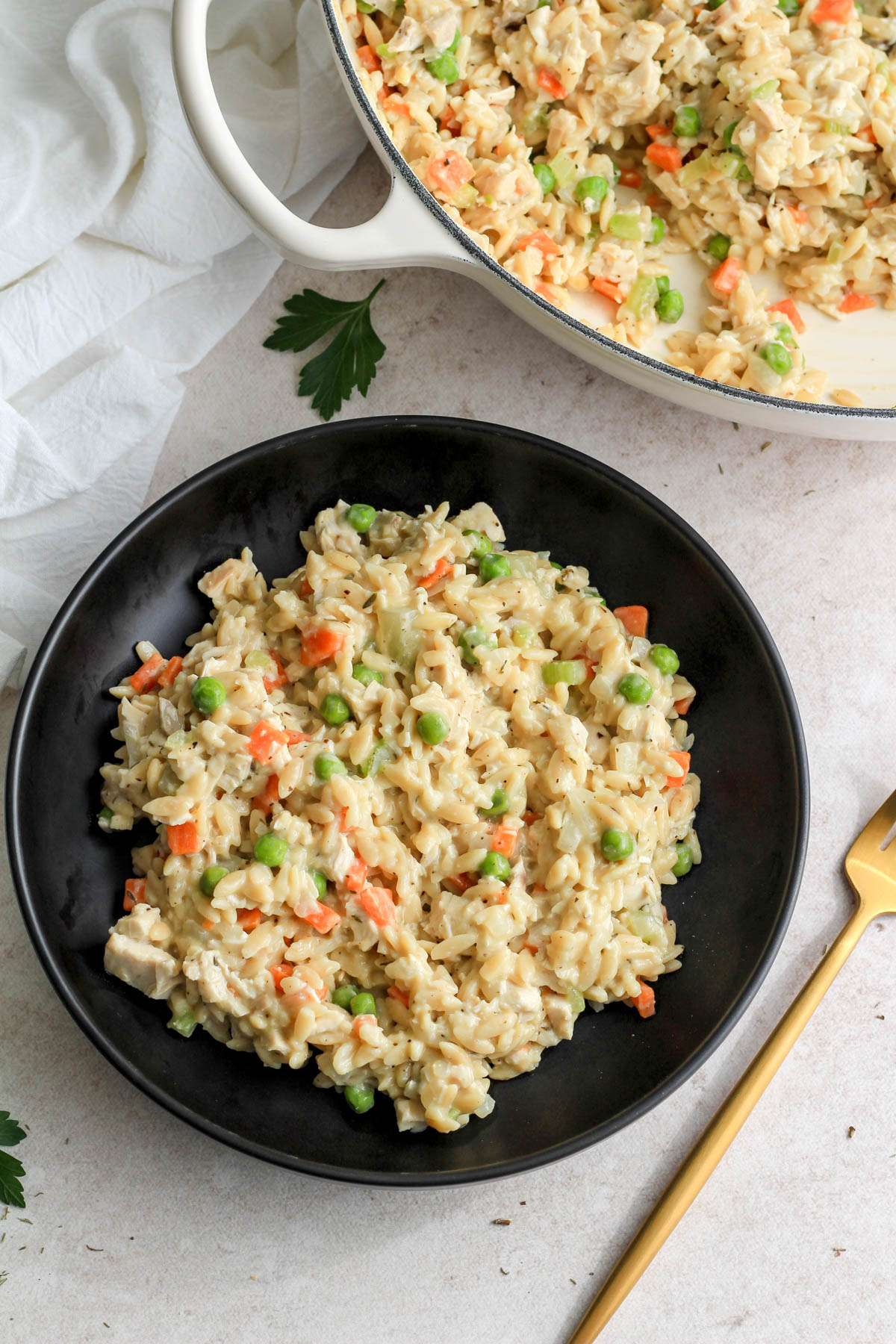 A black bowl of chicken pot pie orzo on a tan counter with a white pan in the back right.
