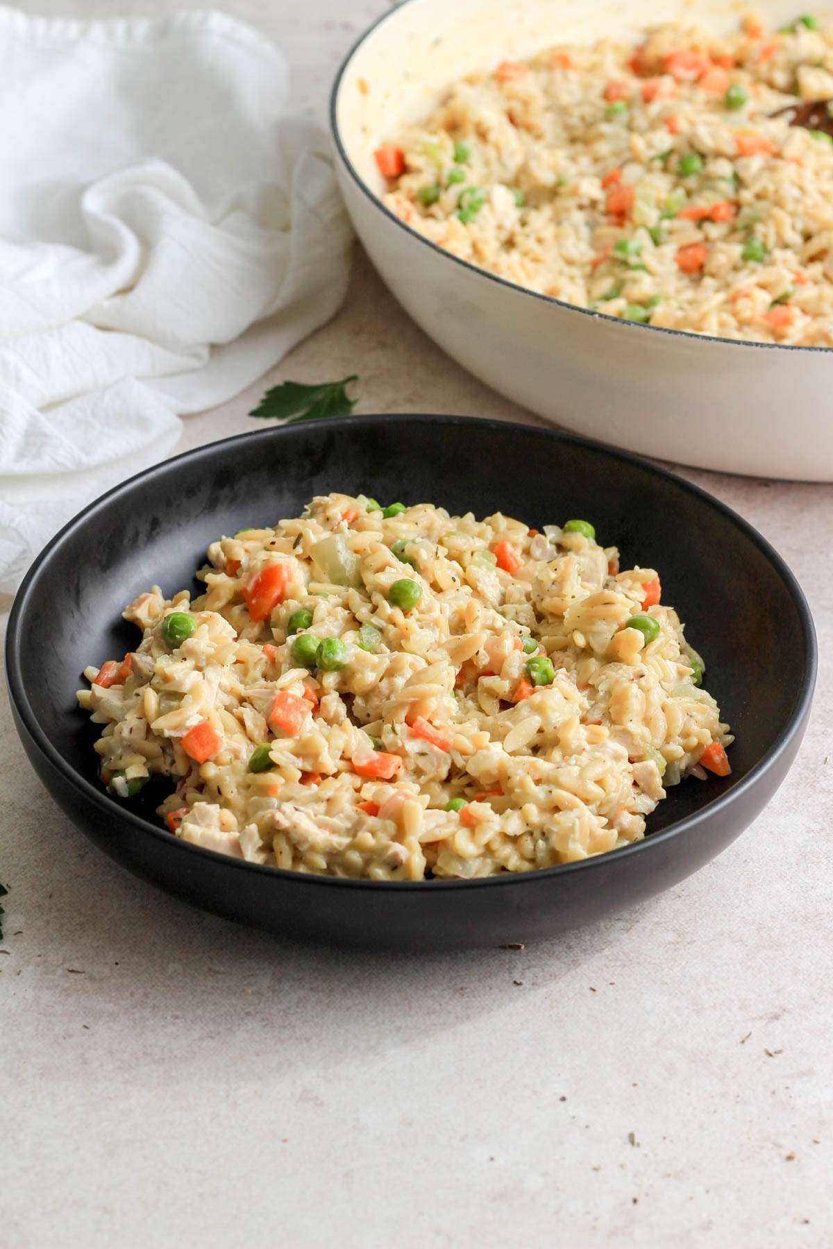 A black bowl with a serving of dairy-free chicken pot pie orzo in front of a white skillet of one pan chicken pot pie orzo.
