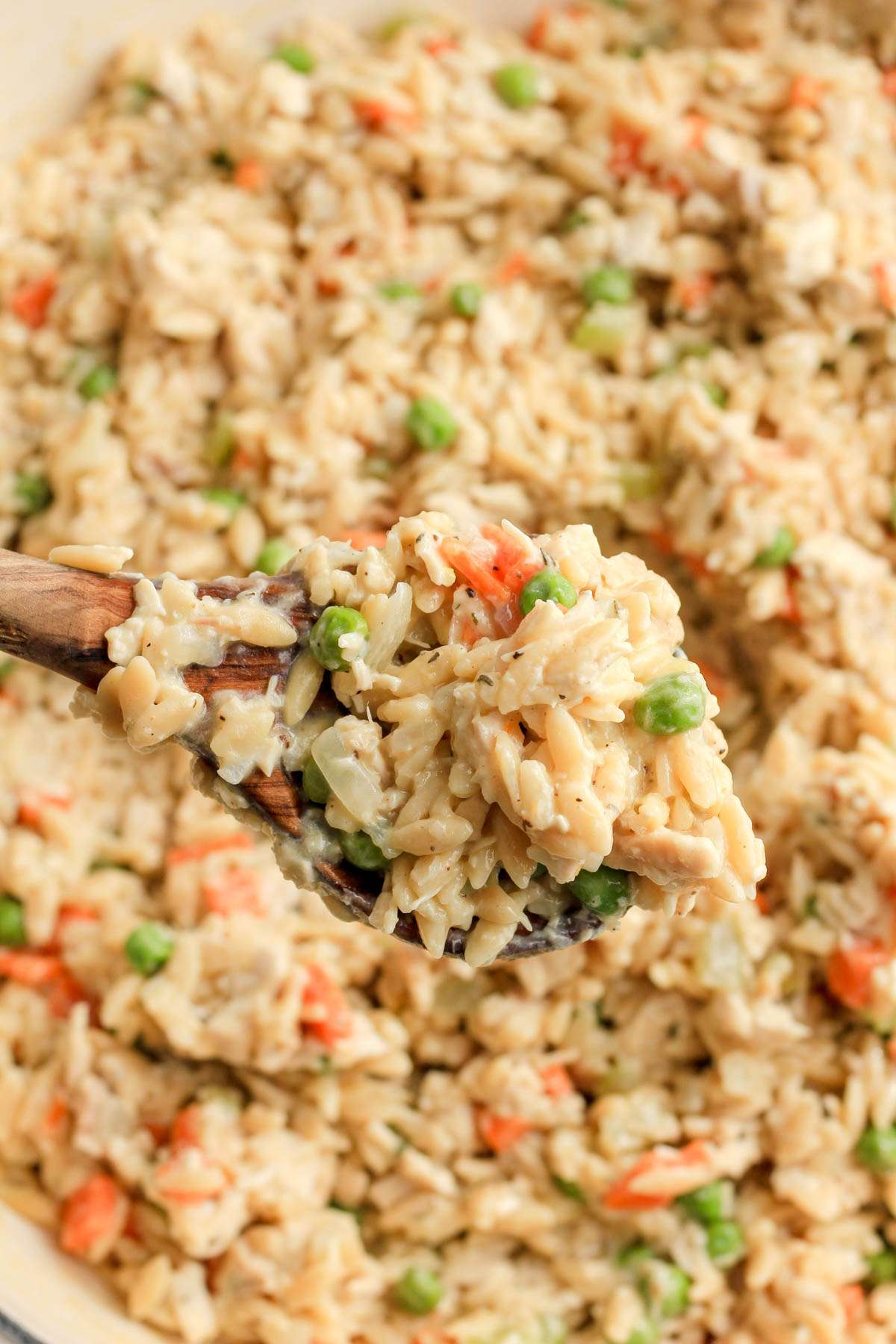 A wooden spoon with a scoop of dairy free chicken pot pie orzo over a white skillet with the one pot orzo dish.